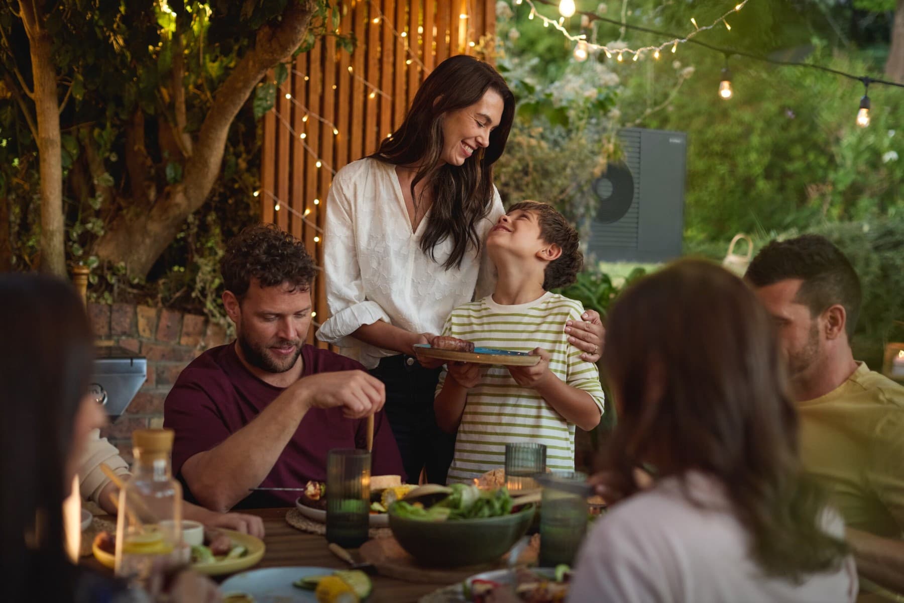 Eine Familie sitzt fröhlich beim Grillen im Garten beisammen. Im Hintergrund steht eine Vaillant aroTHERM plus Wärmepumpe in schwarzgrau.