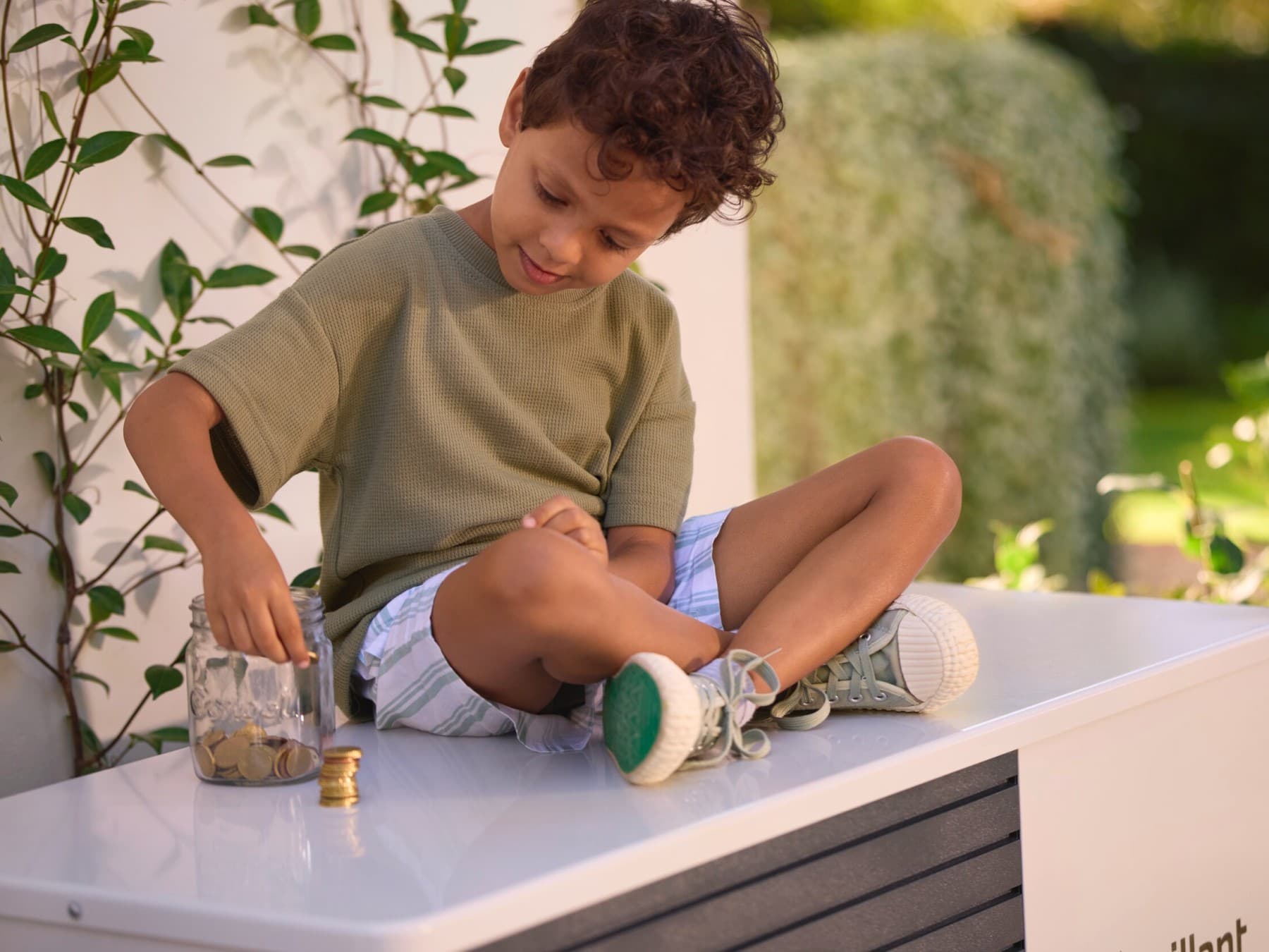 Ein Junge sitzt auf einer Vaillant Wärmepumpe und stapelt Geldmünzen neben einem Einmachglas, in dem weitere Münzen sind.