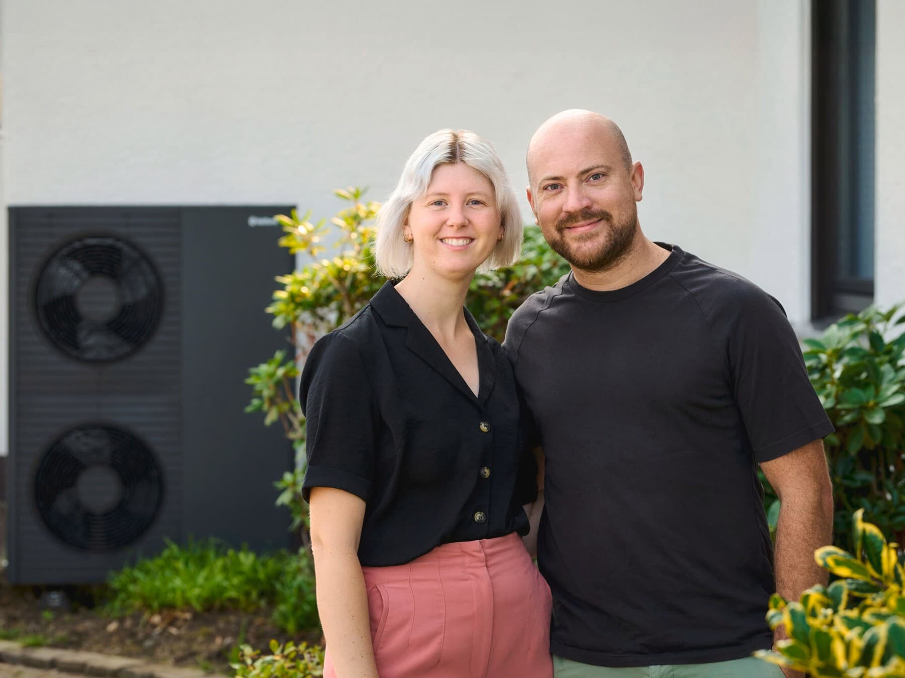 Pia Bondke und Fabian Schiedeck stehen Arm in Arm vor Ihrem Haus. Im Hintergrund ist die Wärmepumpe zu sehen.