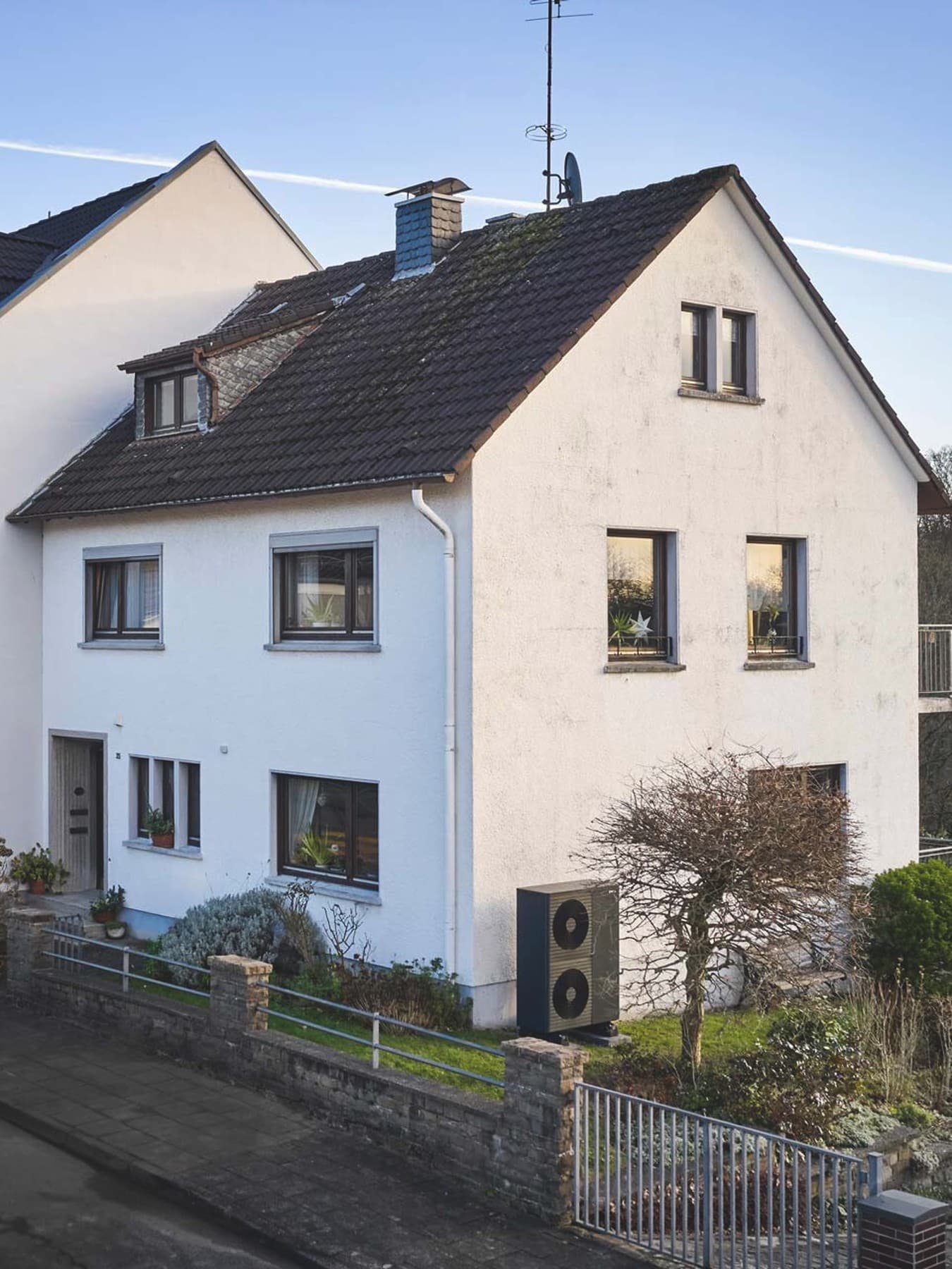 Freistehendes Einfamilienhaus mit hellem Fassadenanstrich und dunklem Dach, umgeben von einem gepflegten Garten mit Rasenflächen. Vor dem Haus befindet sich eine gepflasterte Einfahrt mit geparktem Fahrzeug, im Hintergrund sind weitere Wohnhäuser und eine ländliche Umgebung zu sehen