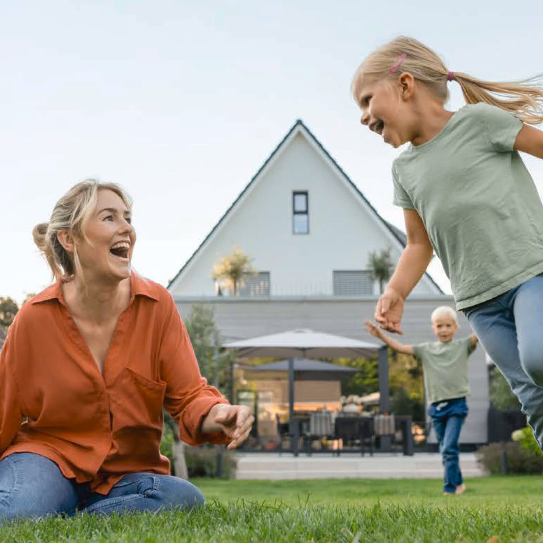 Eine Frau spielt mit zwei Kindern in einem Garten hinter einem Haus.