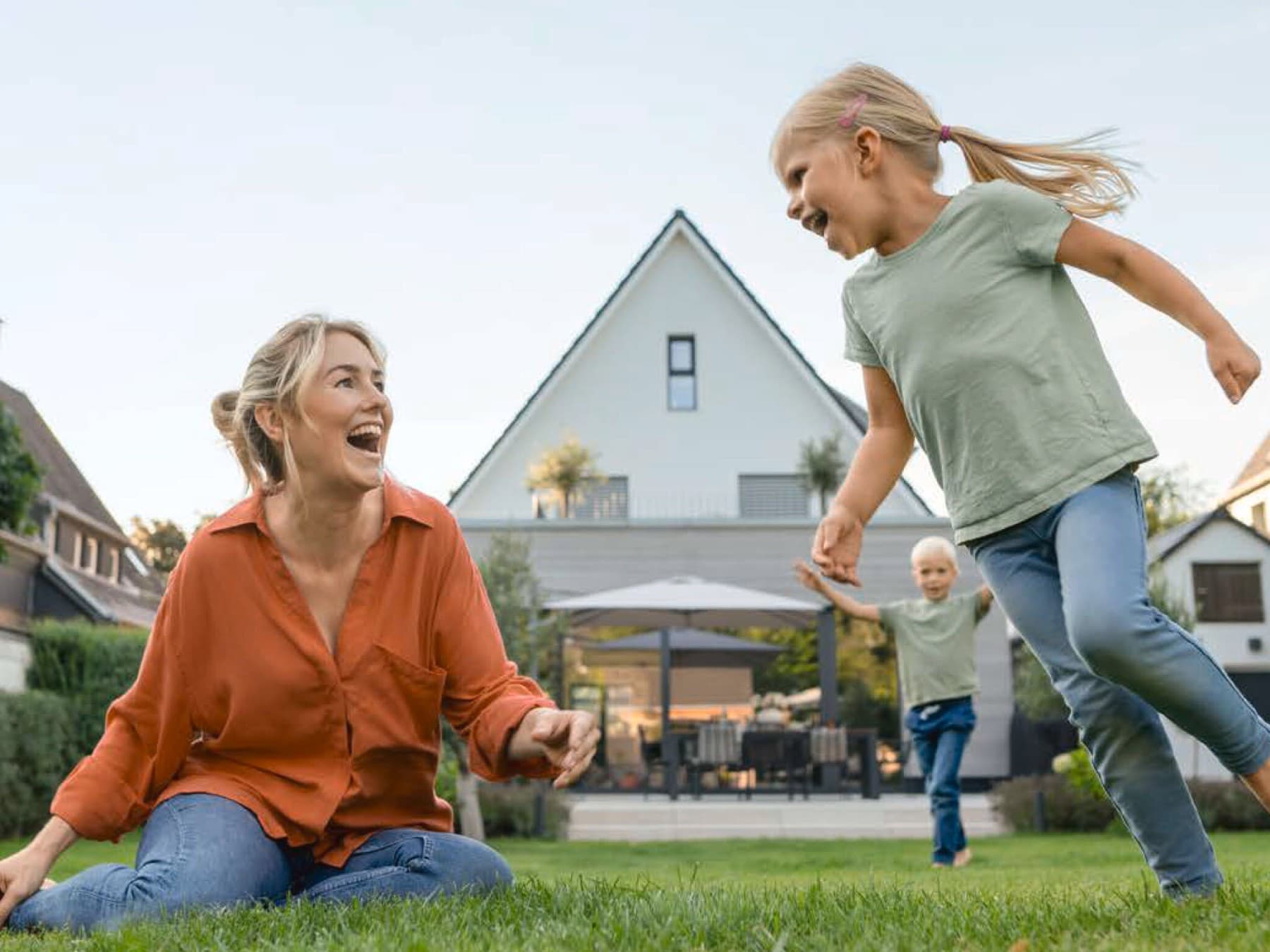 Eine Frau spielt mit zwei Kindern im Garten hinter einem Haus mit Terrasse.