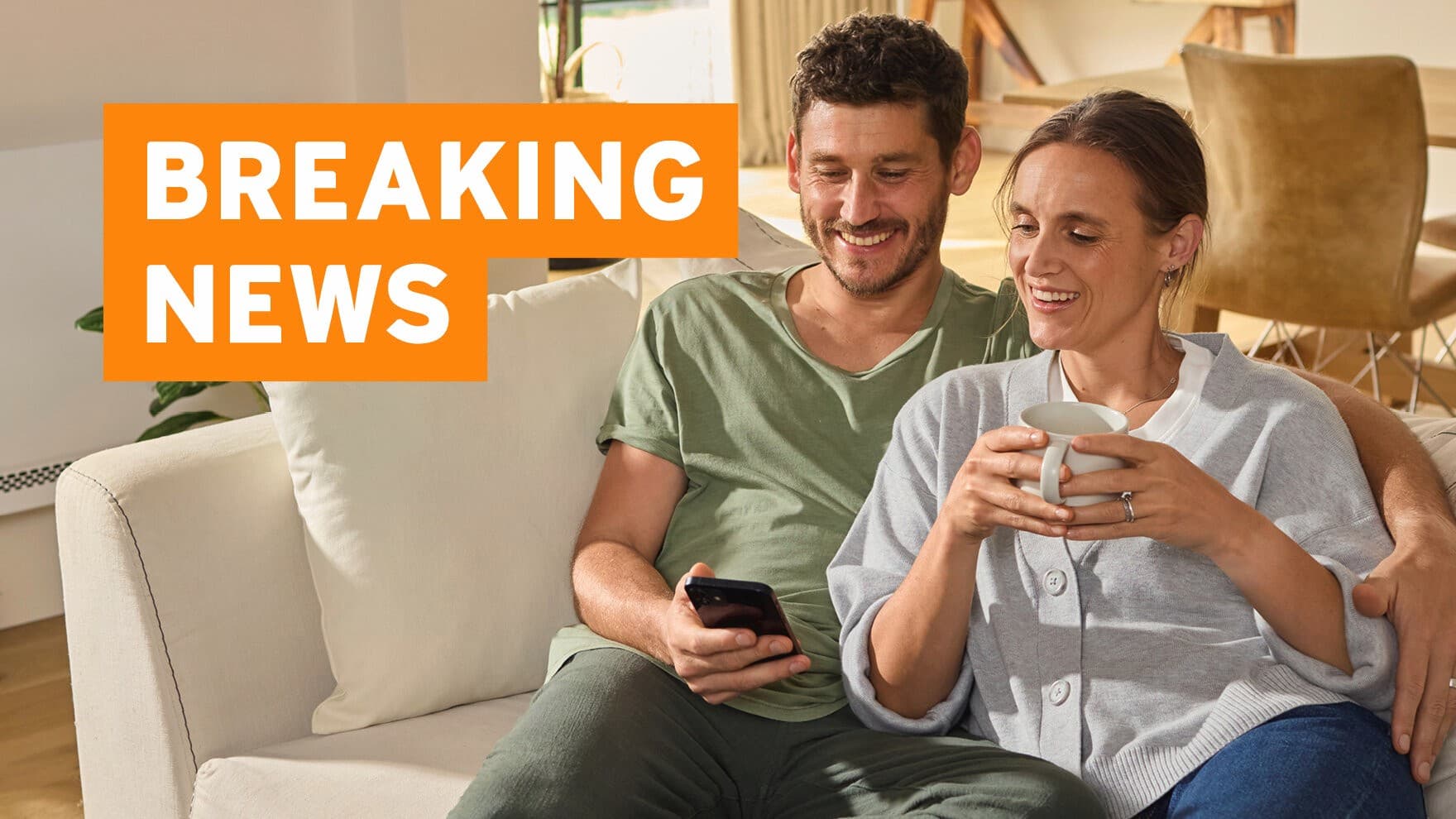 Ein Mann und eine Frau sitzen gemeinsam auf einer hellen Couch und schauen fröhlich auf ein Smartphone, das der Mann in der Hand hält. Die Frau hält eine Tasse in den Händen.