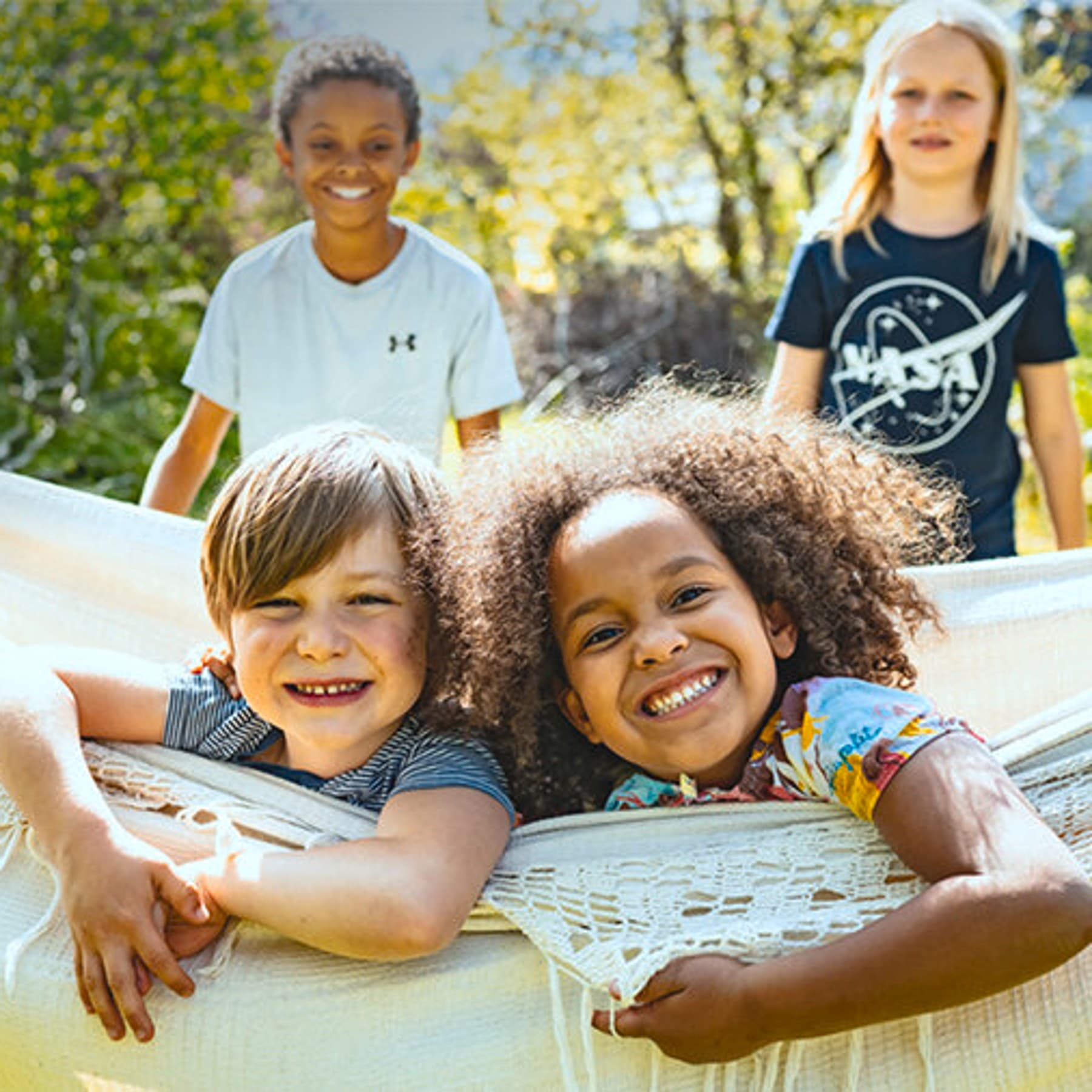 zwei Kinder liegen lachend in einer Hängematte und schauen in die Kamera, im Hintergrund stehen neben der Hängematte zwei weitere fröhliche Kinder.