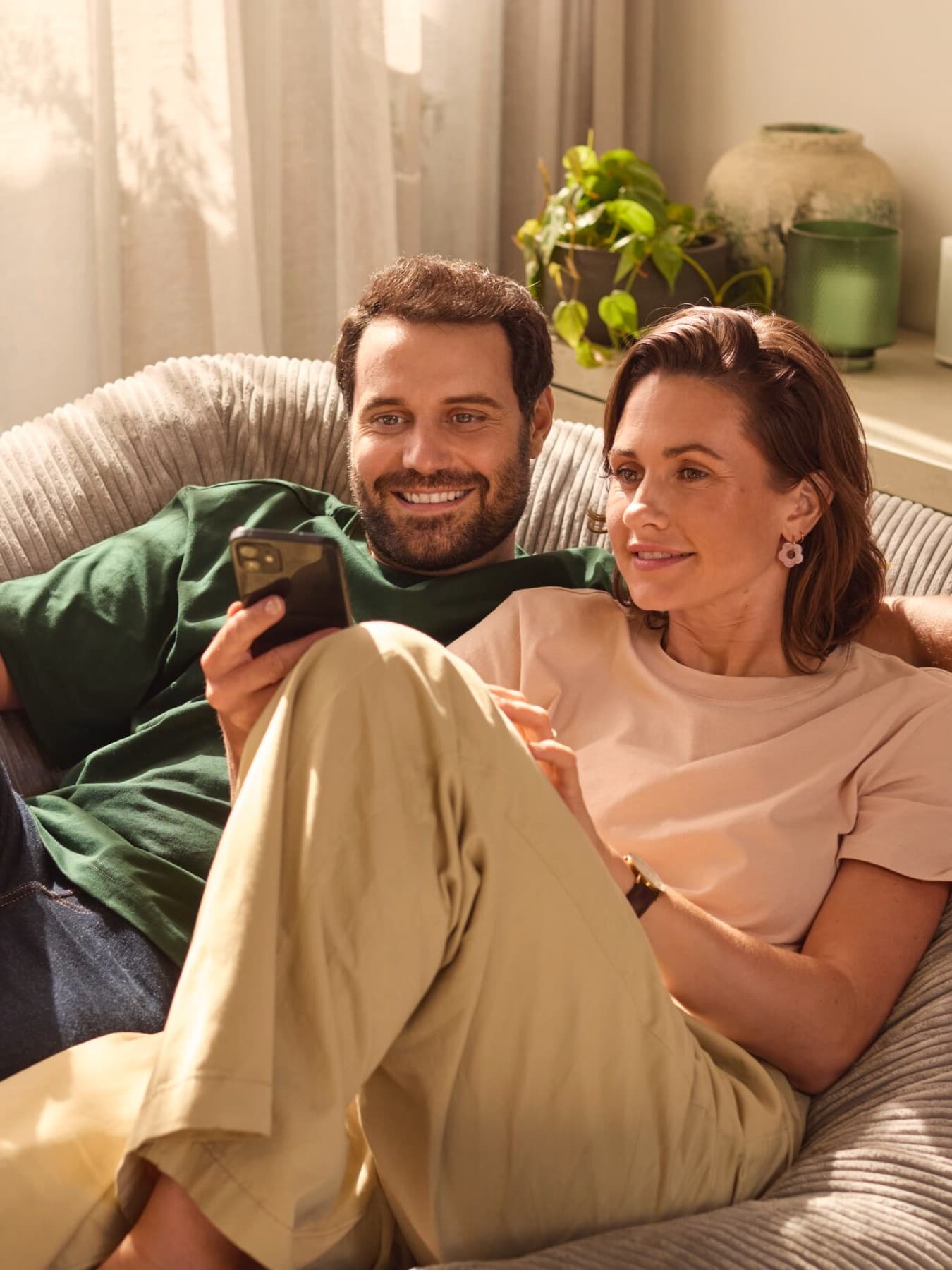 Eine Frau und ein Mann sitzen gemeinsam auf einer Couch und schauen auf ein Smartphone. Der Mann hat der Frau den Arm um die Schulter gelegt und lächelt. Im Hintergrund sieht man eine grüne Pflanze und farblich passende Deko auf einem Sideboard sowie eine weiße Gardine.