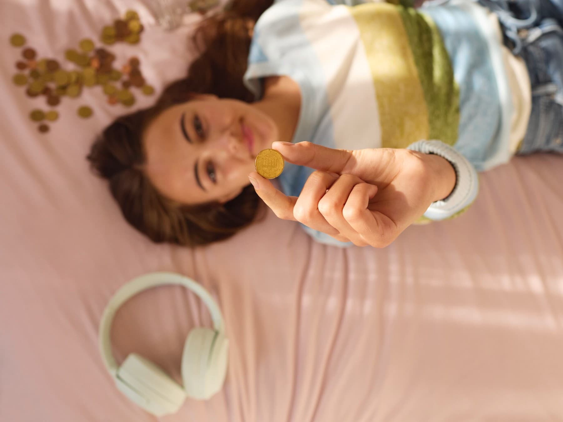 Eine junge Frau liegt auf dem Rücken auf einem weichen Untergrund. Rechts neben ihrem Kopf liegen Kopfhörer, links Geldmünzen. Eine Geldmünzen hält sie hoch in Richtung Kamera.