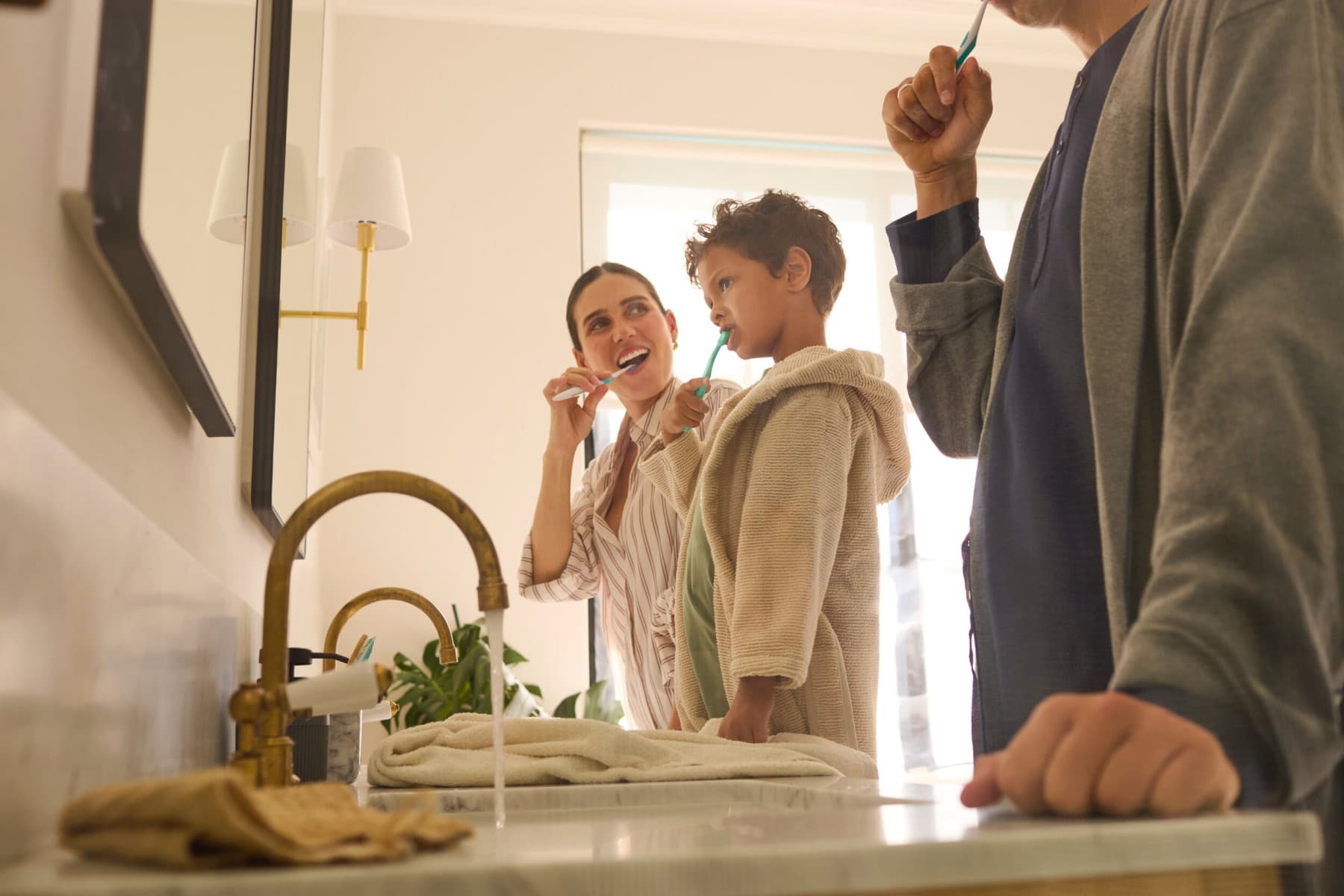 Zwei Erwachsene und ein Kind stehen im Badezimmer vor einem Spiegel und putzen sich die Zähne