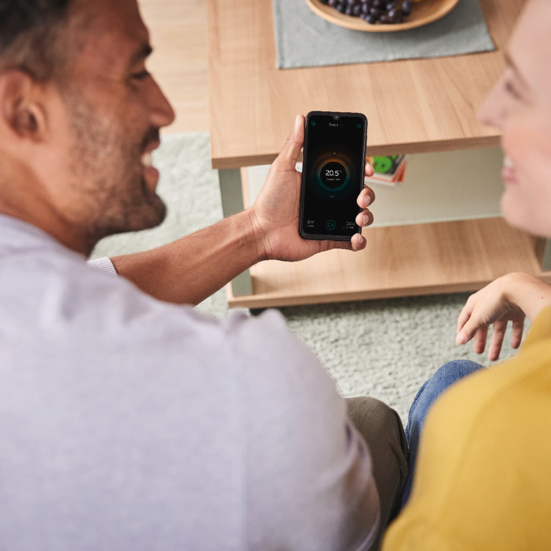 Eine Frau und ein Mann sitzen auf einer Couch, über ihre Schulter hinweg sieht man auf ein Smartphone-Display, auf dem die myVAIILANT App geöffnet ist.