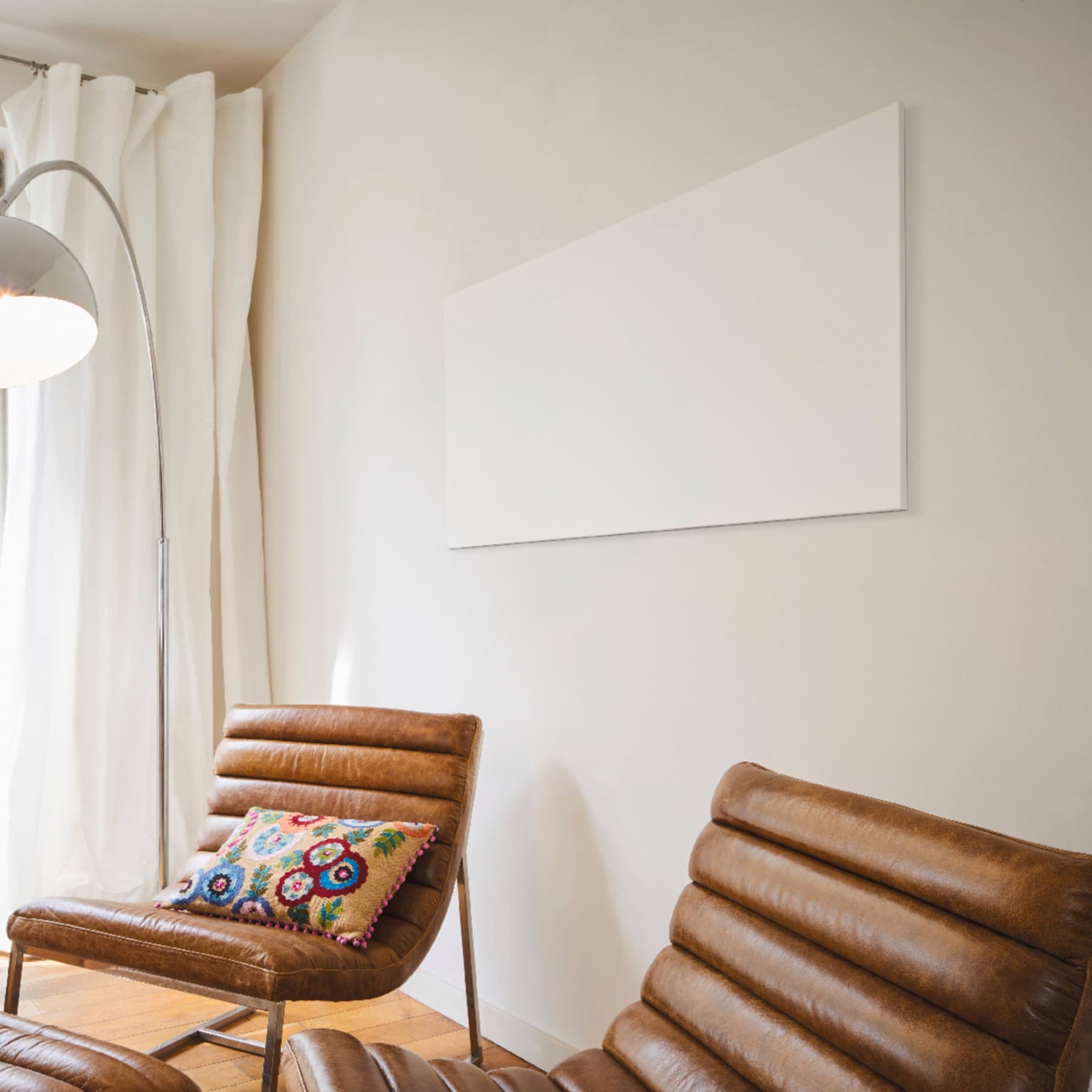 Eine Infrarotheizung hängt an einer Wand über zwei braunen Lederstühlen, die in einem Wohnzimmer vor der Wand stehen. Im Hintergrund befindet sich eine angeschaltete Stehlampe sowie ein bodenhohes Fenster mit Blick in einen Garten.