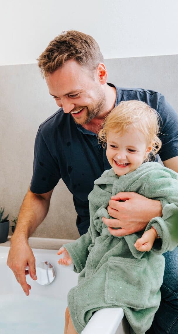 Ein Mann sitzt mit einem Kind im Arm auf dem Rand einer Badewanne und schaut zu, wie das Wasser einläuft.