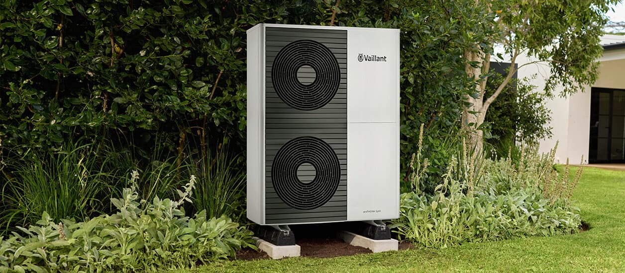 Eine Vaillant Luft-Wasser-Wärmepumpe aroTHERM split steht in einem Garten vor Büschen.