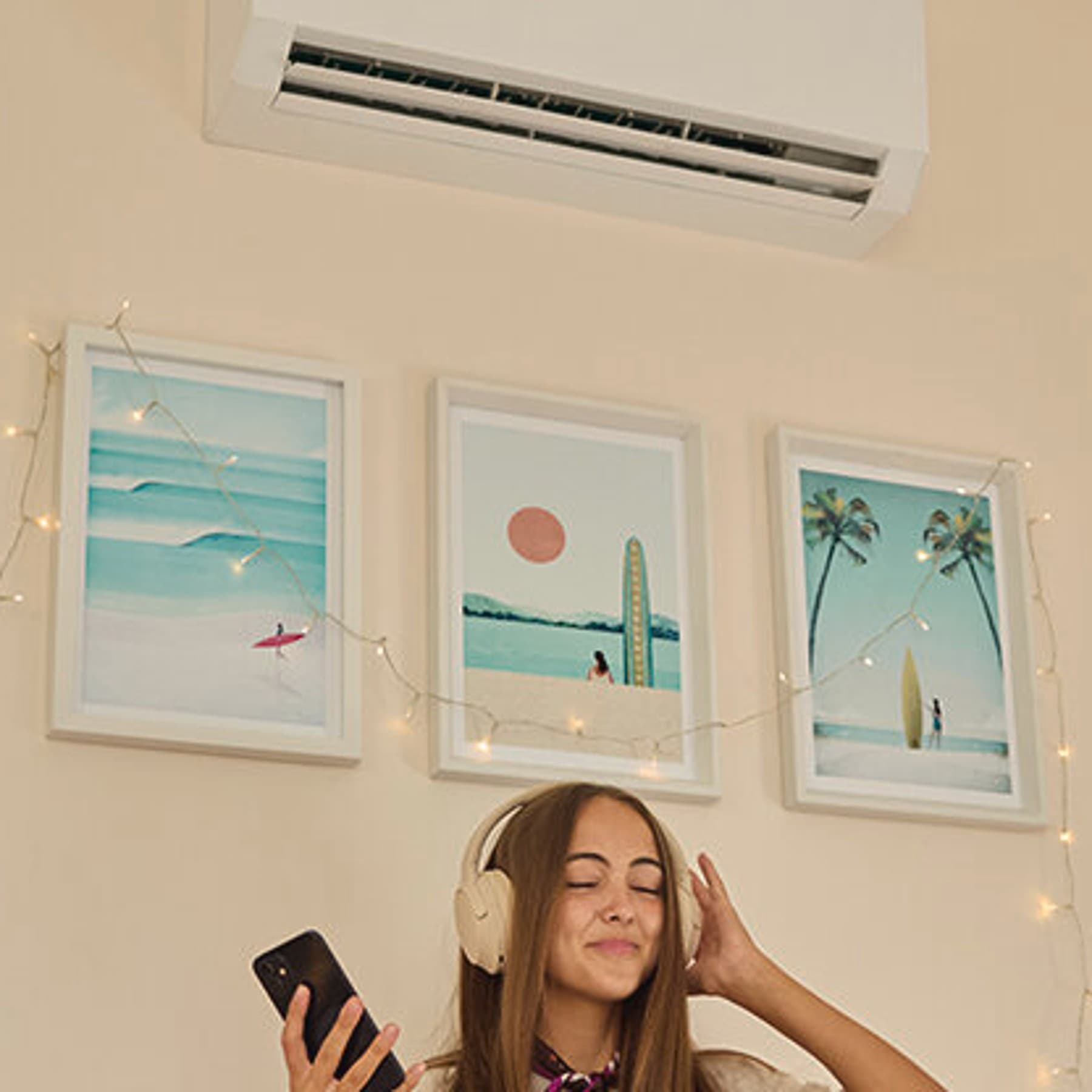 Eine junge Frau sitzt mit Kopfhörern auf dem Kopf und einem Smartphone in der Hand auf der Couch. An der Wand hinter ihr hängen Bilder und eine Vaillant Klimaanlage.