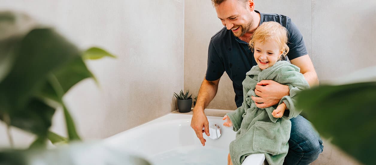 Ein Mann sitzt mit einem Kind in Bademantel auf einem Badewannenrand, während das Wasser einläuft.
