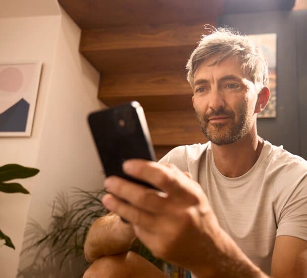Ein Mann sitzt in einem Raum mit einer Treppe im Hintergrund. Er hält ein Smartphone in der linken Hand, auf das er schaut.
