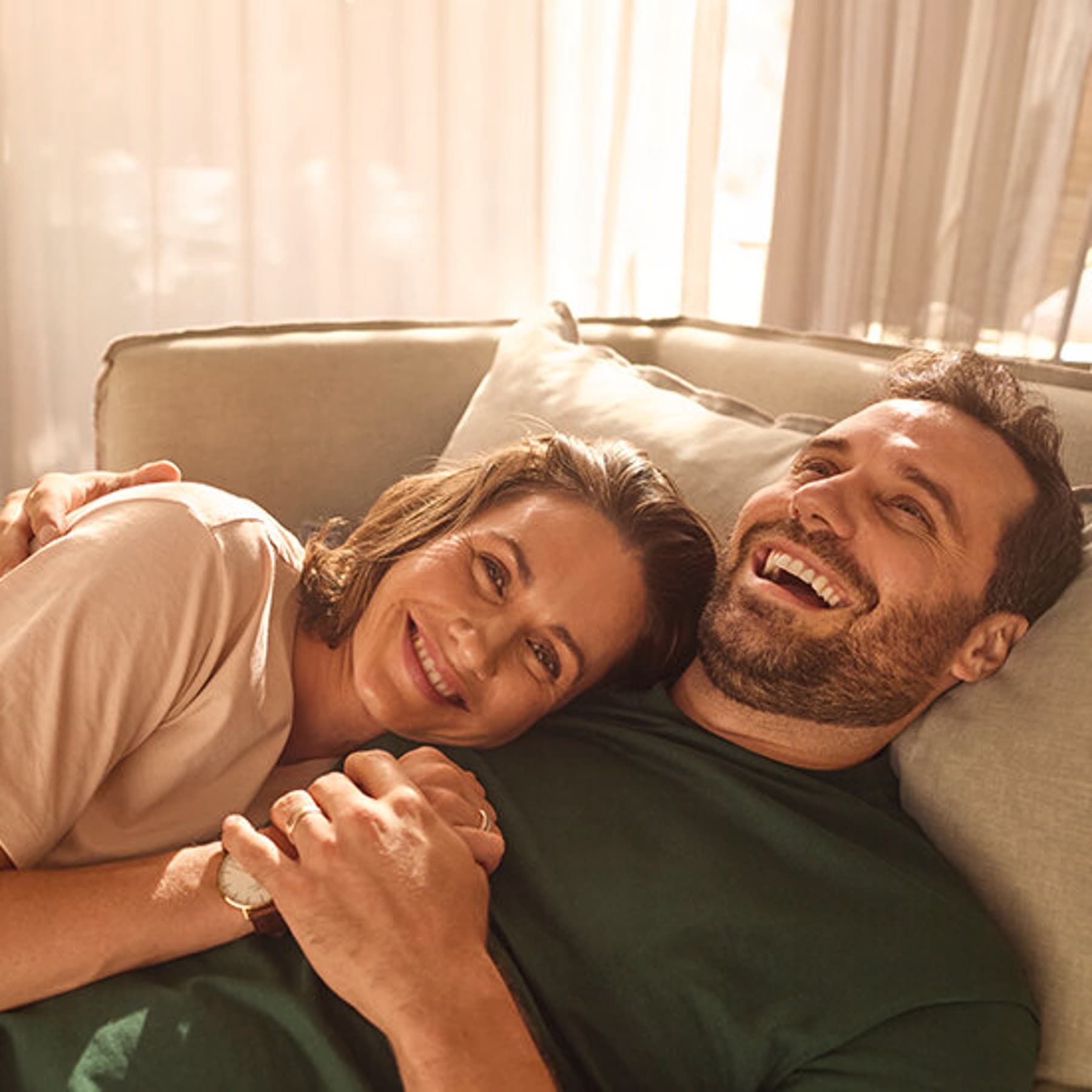 Ein Mann liegt mit einer Frau im Arm fröhlich lachend auf einem Sofa.