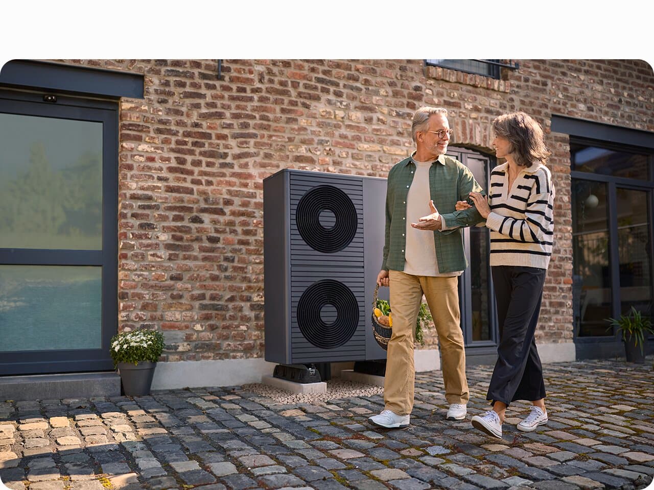 Eine Frau und ein Mann gehen Arm in Arm über einen Kopfsteinpflasterweg. Im Hintergrunnd ist ein Backsteingebäude zu sehen, vor dem eine Wärmepumpe von Vaillant neben dem Kopfsteinweg steht.
