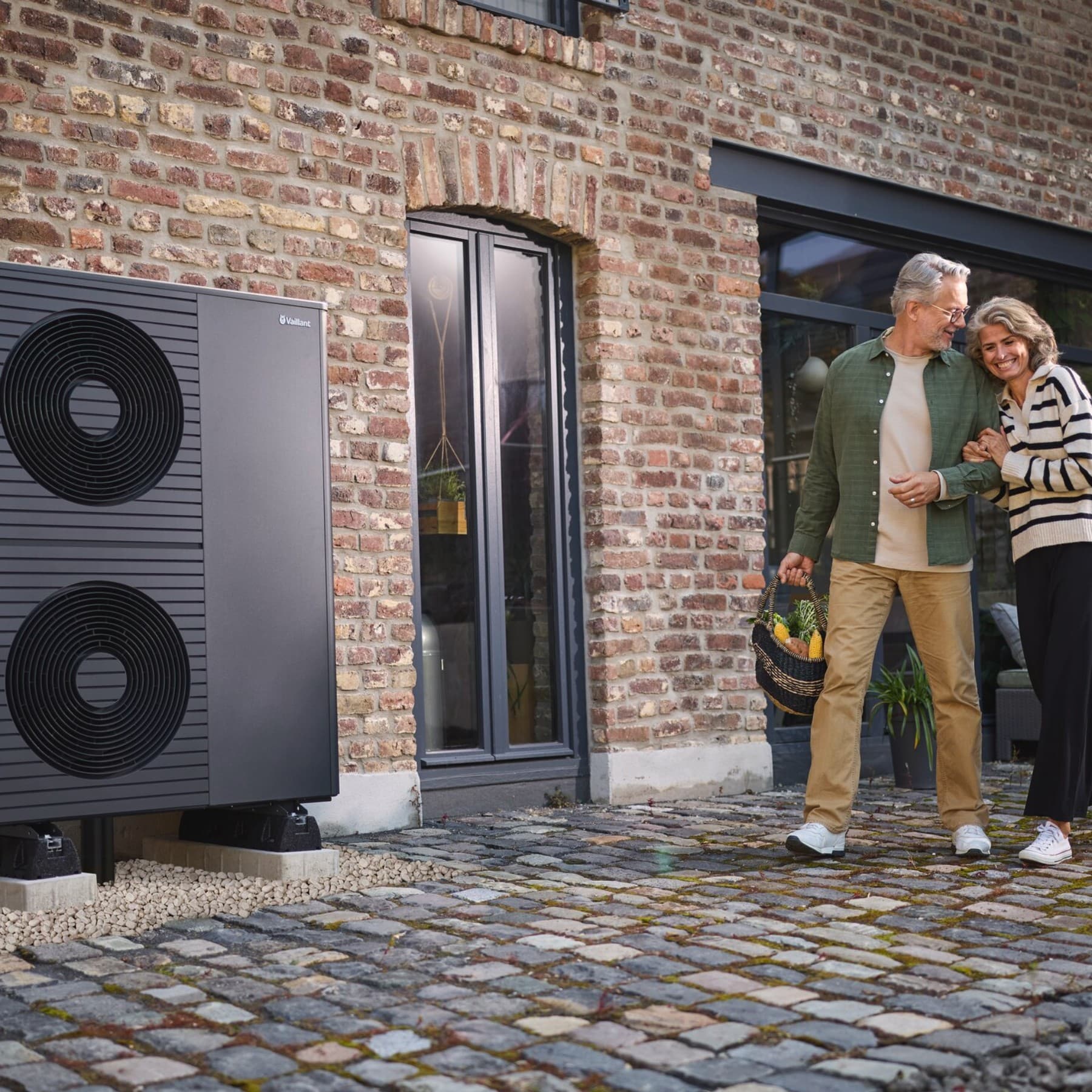 Ein Mann und eine Frau mit weiß-grauen Haaren gehen über einen gepflasterten Weg an an einem Backsteingebäude vorbei. Der Mann hält einen Einkaufskorb in der Hand, die Frau hakt sich bei ihm ein. Am Rand des Weges steht eine Vaillant Wärmepumpe mit zwei Ventilatoren auf einem Kiesbett vor dem Gebäude.