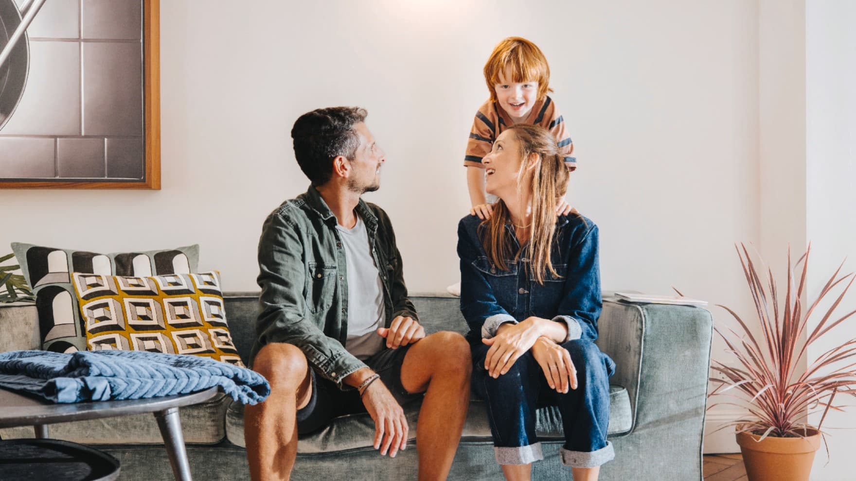 Ein mann und eine Frau sitzen in einem gemütlichen Wohnzimmer auf ihrer Couch, während ein Kind hinter ihnen mit ihnen spielt.