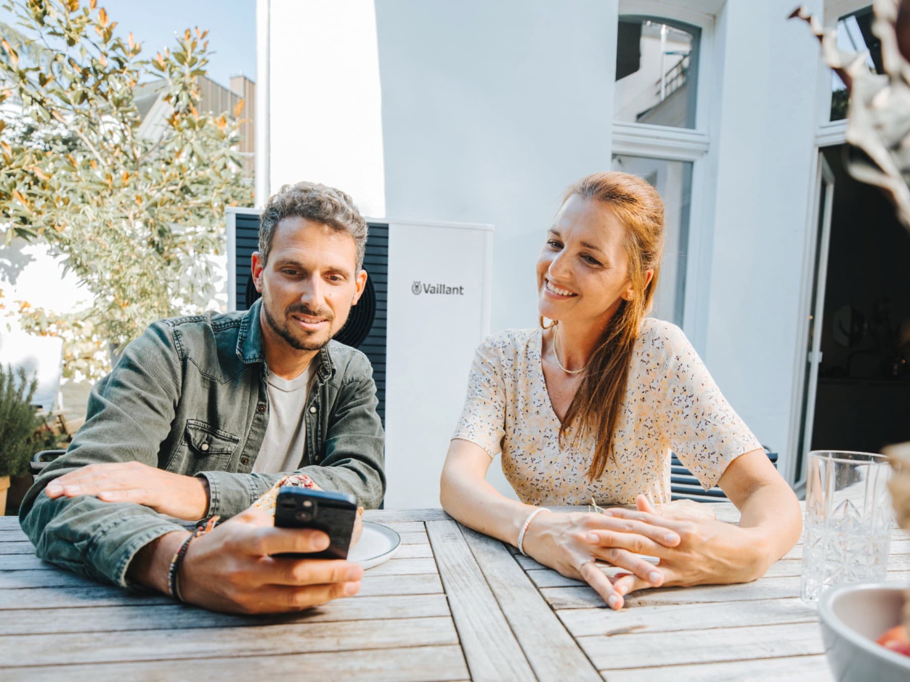 Pärchen am Gartentisch sitzend mit einem Handy in der Hand. Im Hintergrund ist eine Vaillant Wärmepumpe zu sehen.