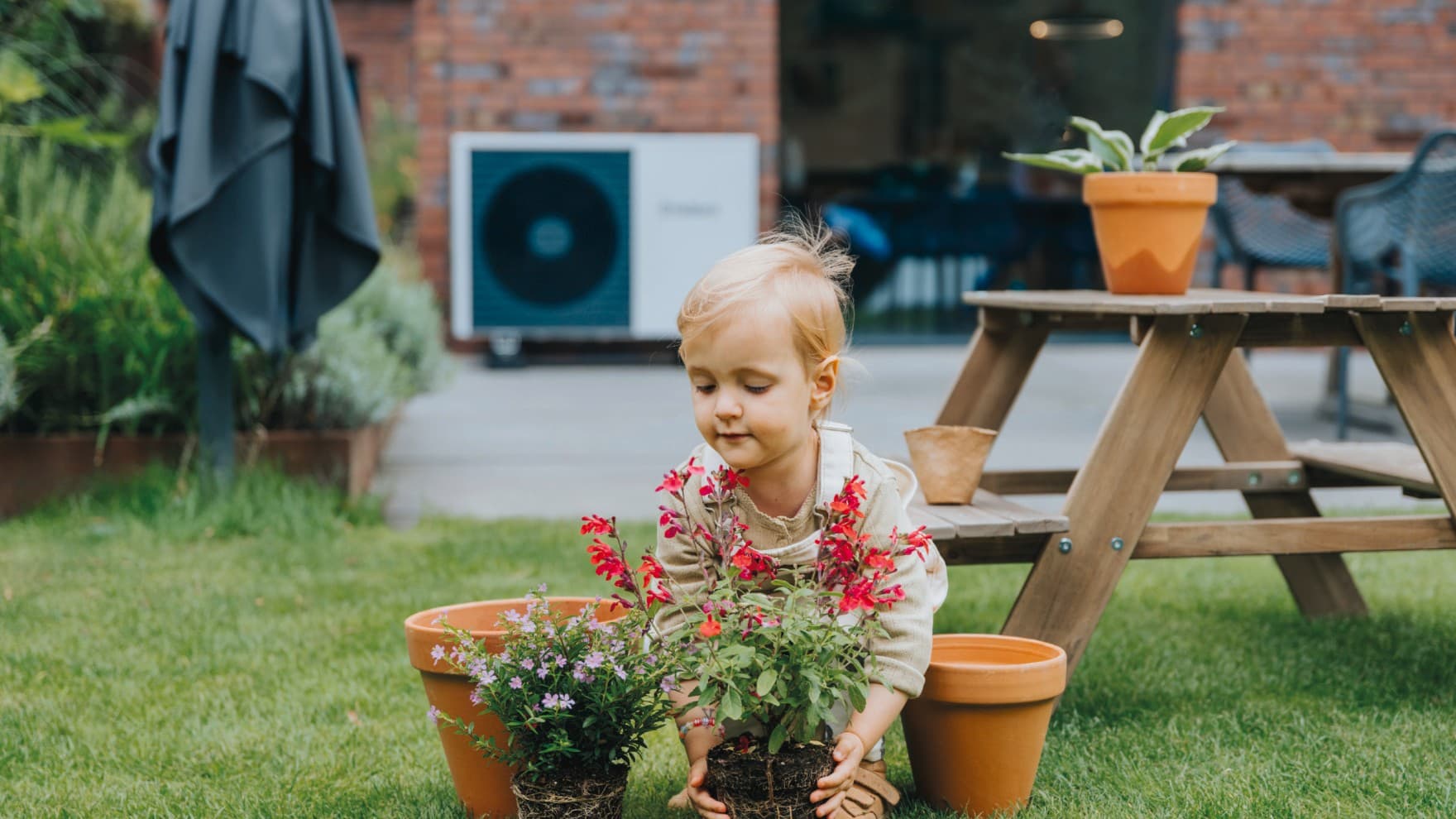 Ein Kind pflanzt im Garten Blumen in einen Topf. Im Hintergrund steht eine Vaillant Wärmepumpe an der Hauswand.