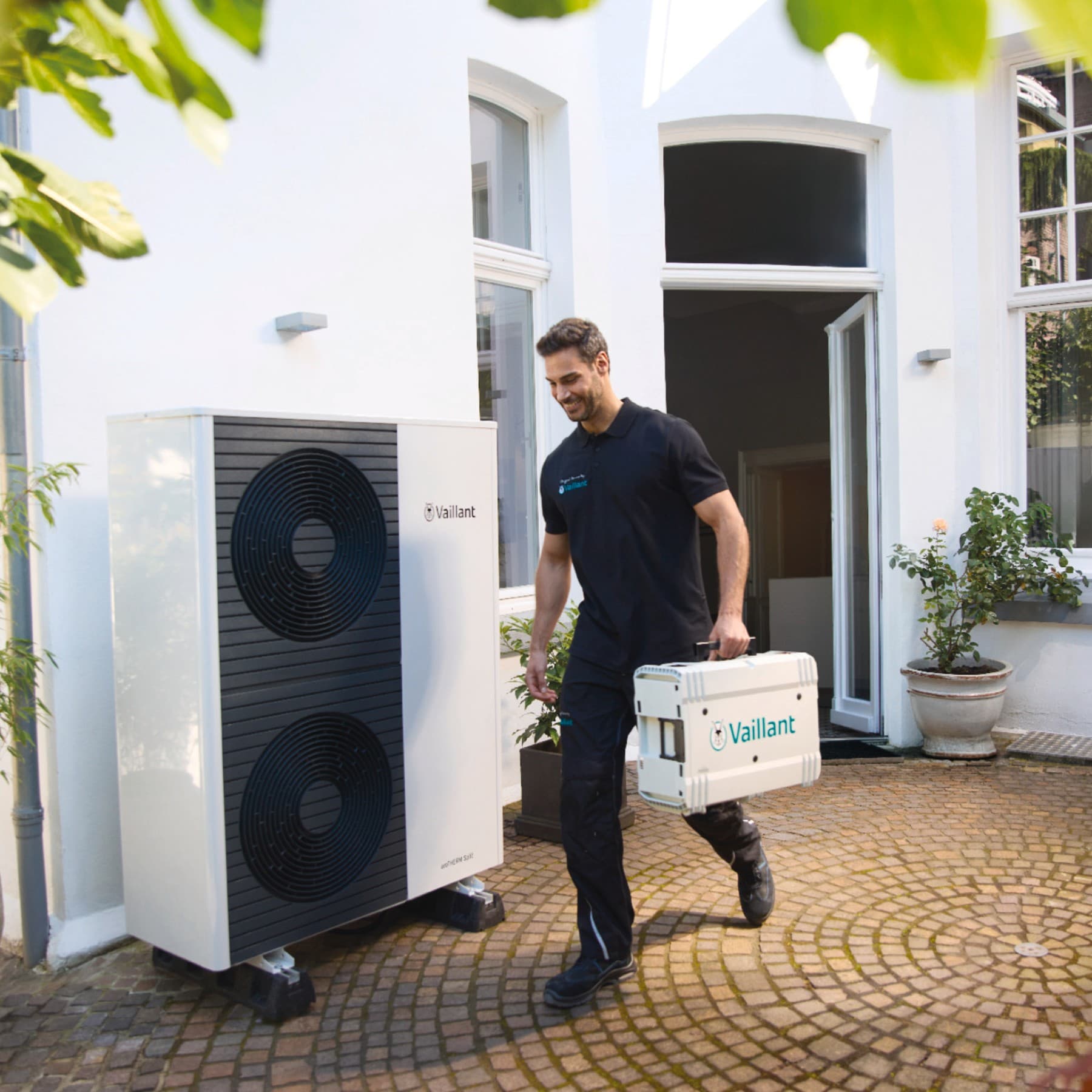Ein Vaillant Techniker geht mit einem Werkzeugkoffer an einer Vaillant Wärmepumpe vorbei, die vor einem Haus steht.