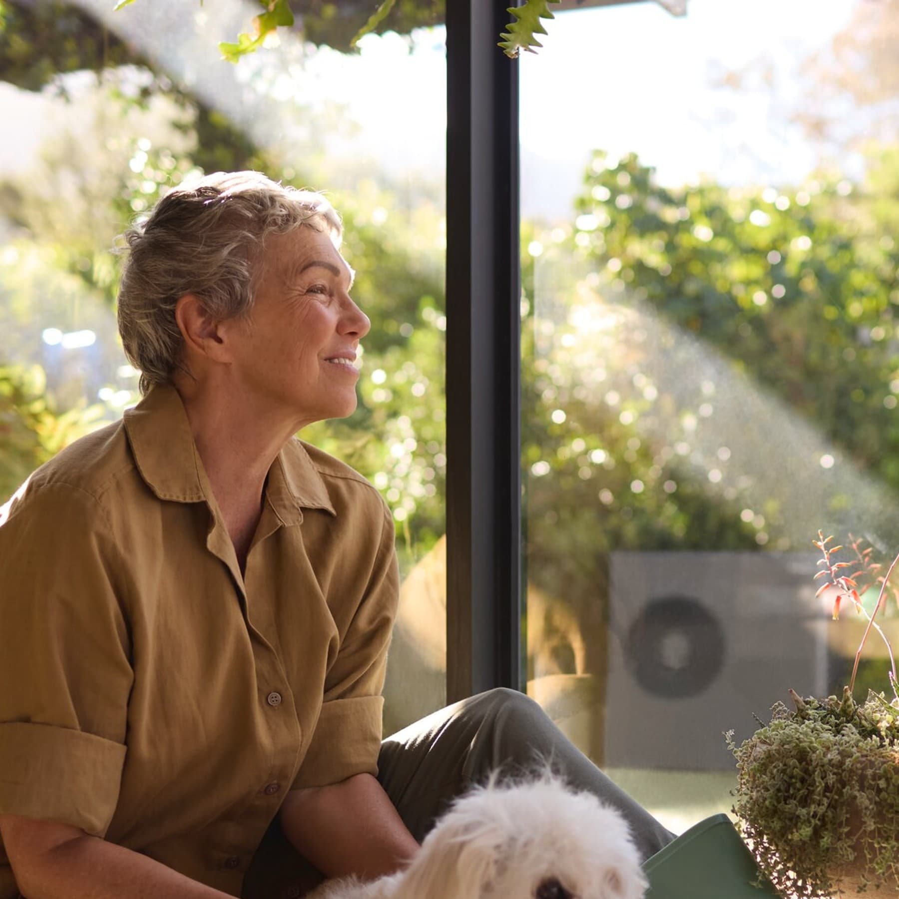 Eine Frau sitzt mit einem weißen Hund zwischen Topfpflanzen. Im Hintergrund sieht man eine schwarzgraue Vaillant Wärmepumpe.