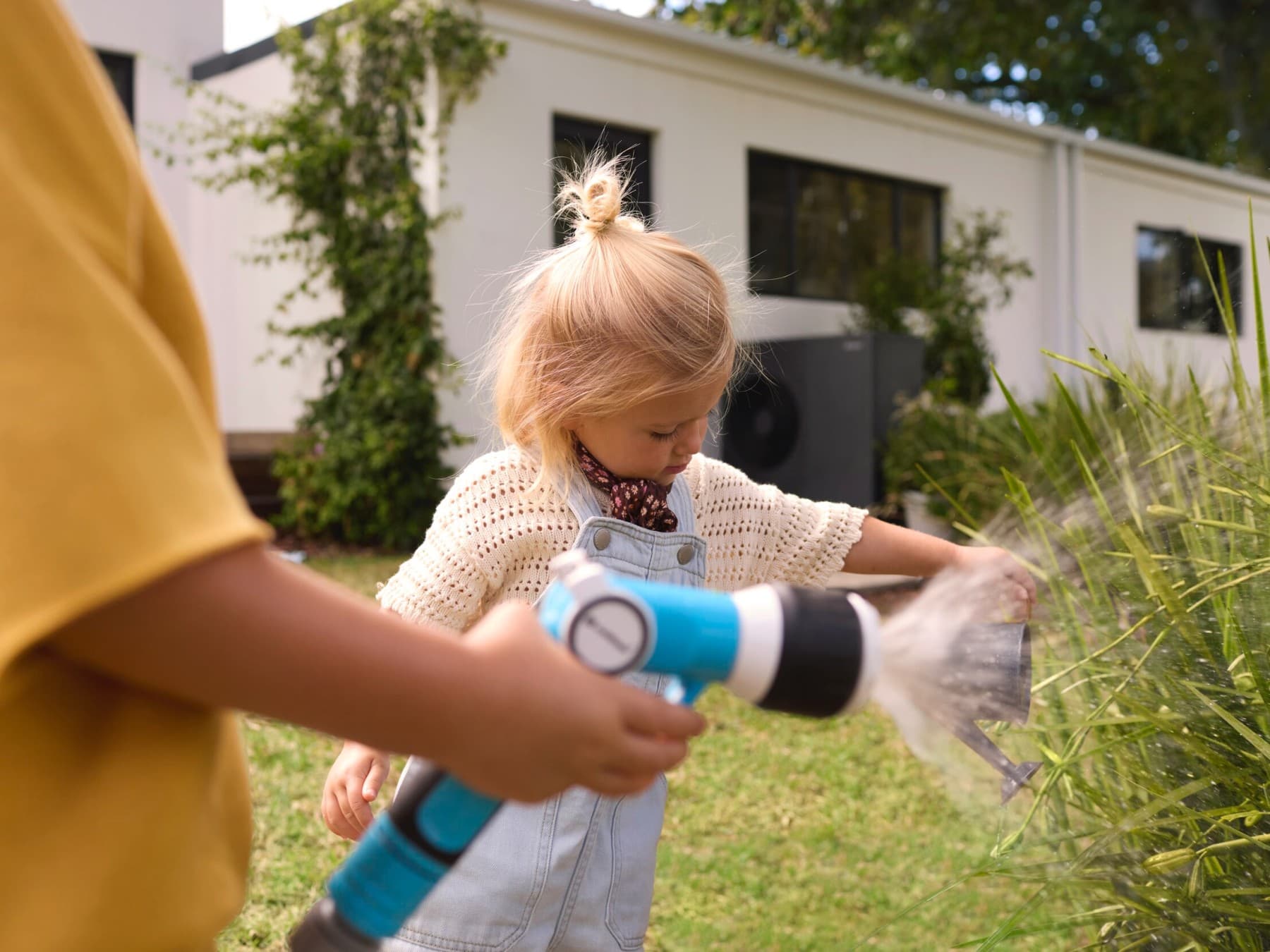 Ein Mensch gießt mit einem Wasserschlauch einen Busch im Garten, daneben steht ein Kind und schaut zu. Im Hintergund steht eine Vaillant Wärmepumpe an der Hauswand.