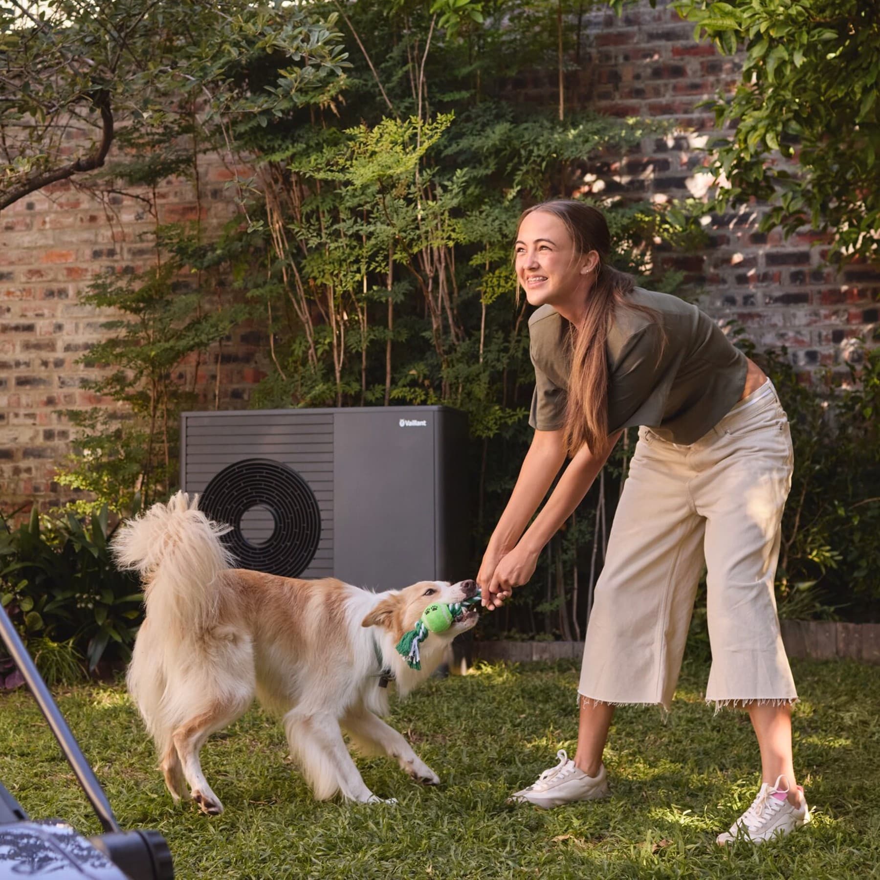 Eine Luftwärmepumpe steht im Garten. Davor spielt ein Mädchen mit einem Hund auf der Wiese.
