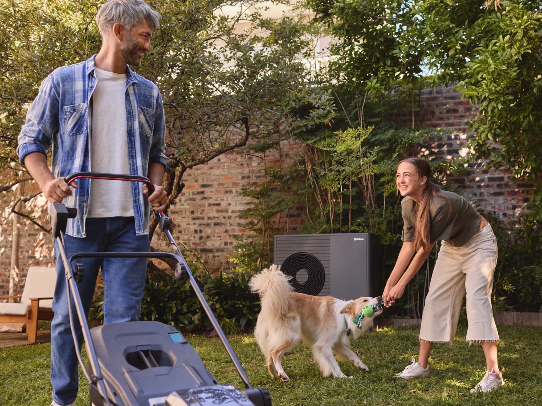 Eine Luftwärmepumpe steht im Garten. Davor spielt ein Mädchen mit einem Hund auf der Wiese.