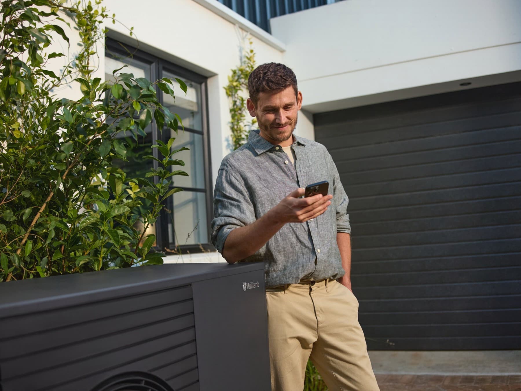 Mann steht mit Smartphone in der Hand neben einer Vaillant Wärmepumpe aroTHERM plus vor einem Haus