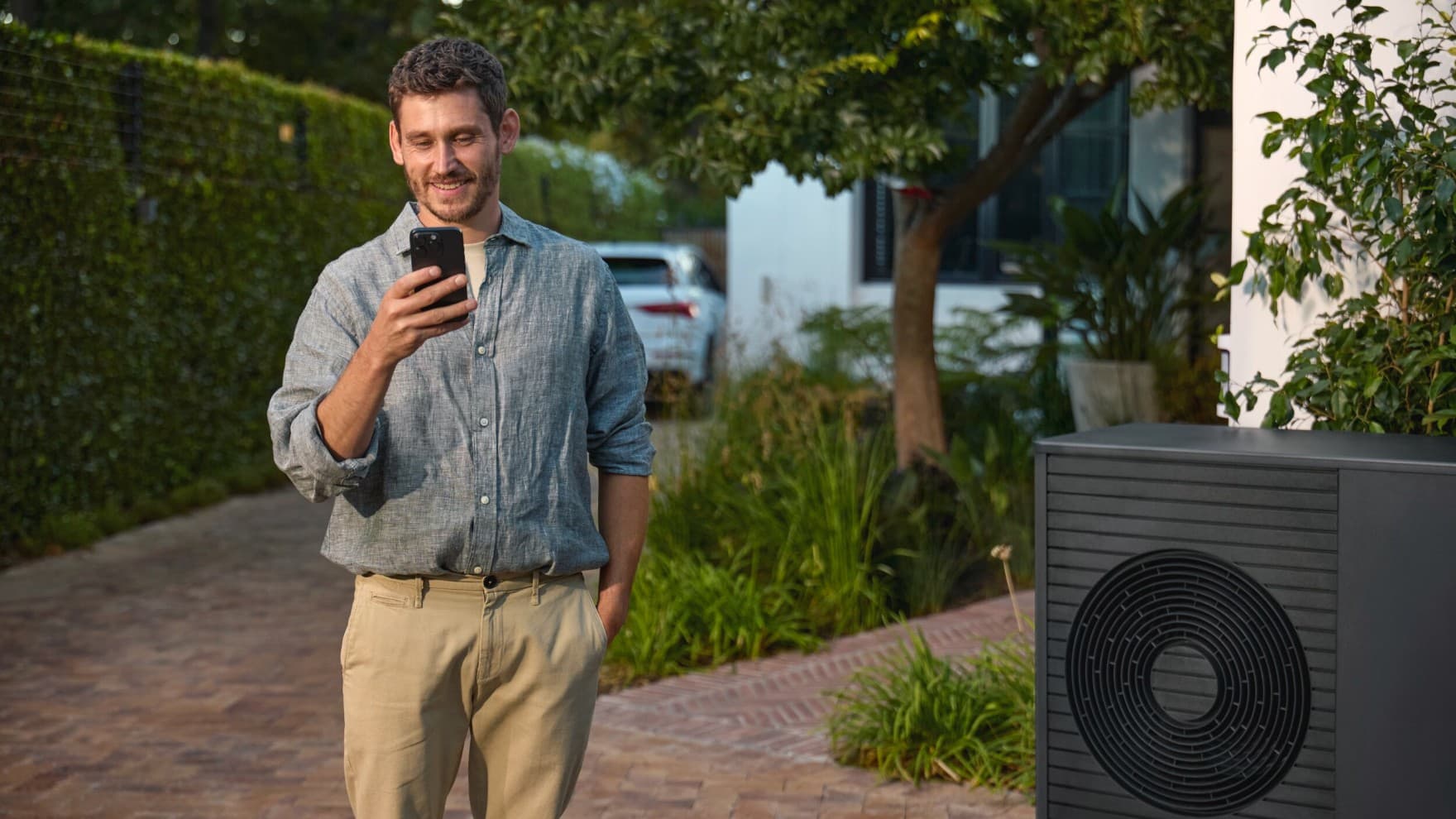 Mann mit Smartphone in der Hand steht neben einer Wärmepumpe aroTHERM plus vor einem Haus.