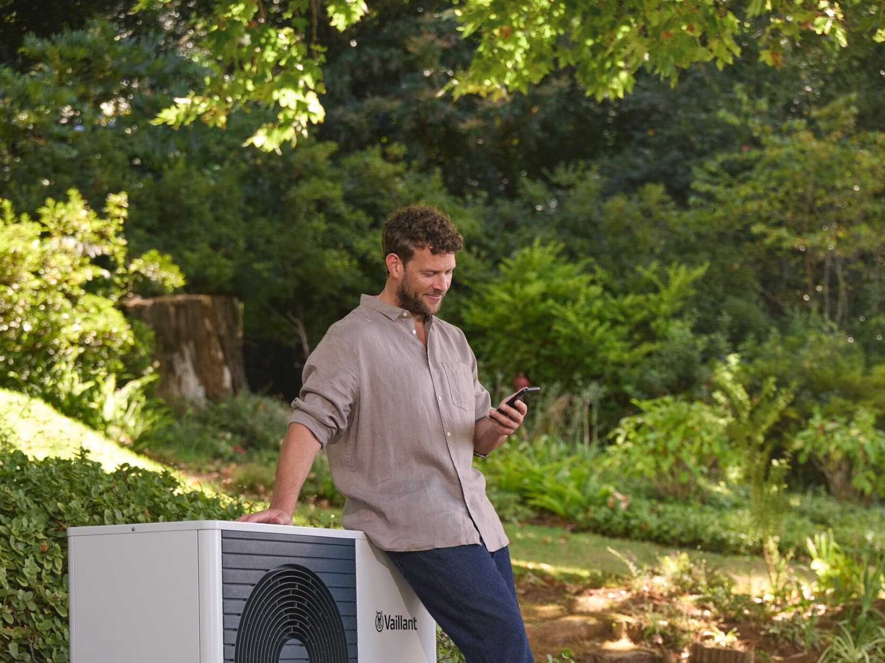 Ein Mann lehnt an einer Vaillant Wärmepumpe, die in einem grünen Garten steht, und schaut auf sein Smartphone.