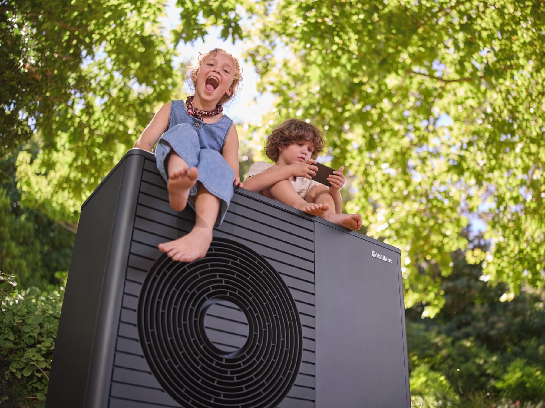 Zwei Kinder, die auf einer schwarzgrauen Vaillant Wärmepumpe sitzen