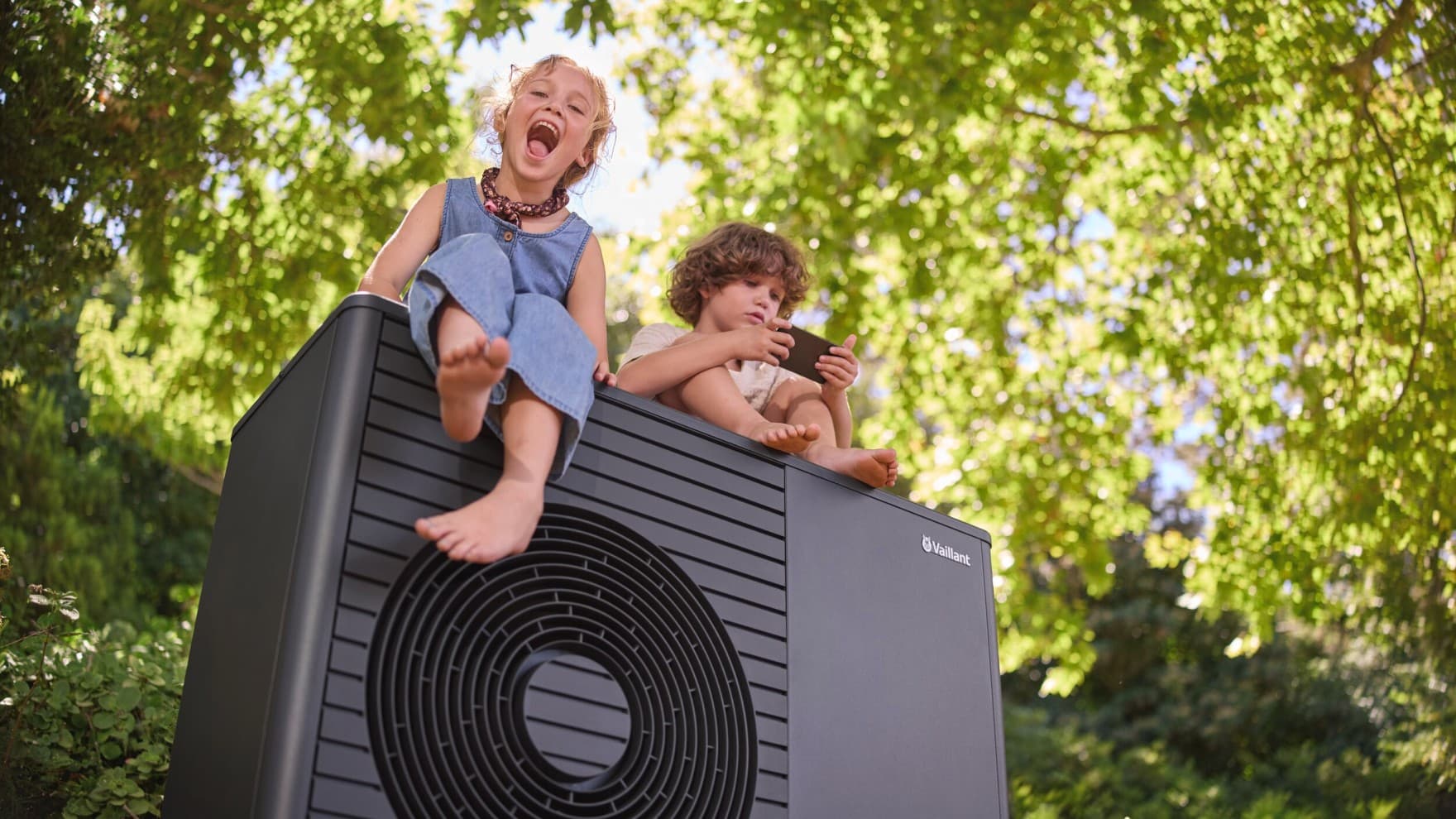 Zwei Kinder sitzen auf einer aroTHERM plus Wärmepumpe im Garten unter Bäumen