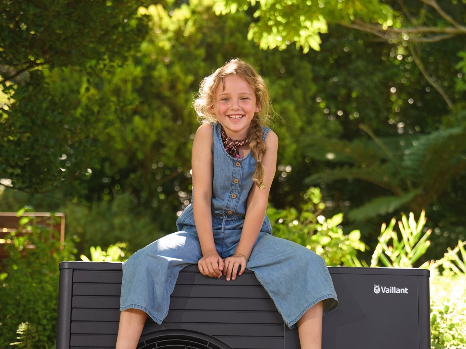 Lachendes Mädchen sitzt auf der Außeneinheit der Vaillant Luft-Wasser-Wärmepumpe aroTHERM plus im Sommergarten