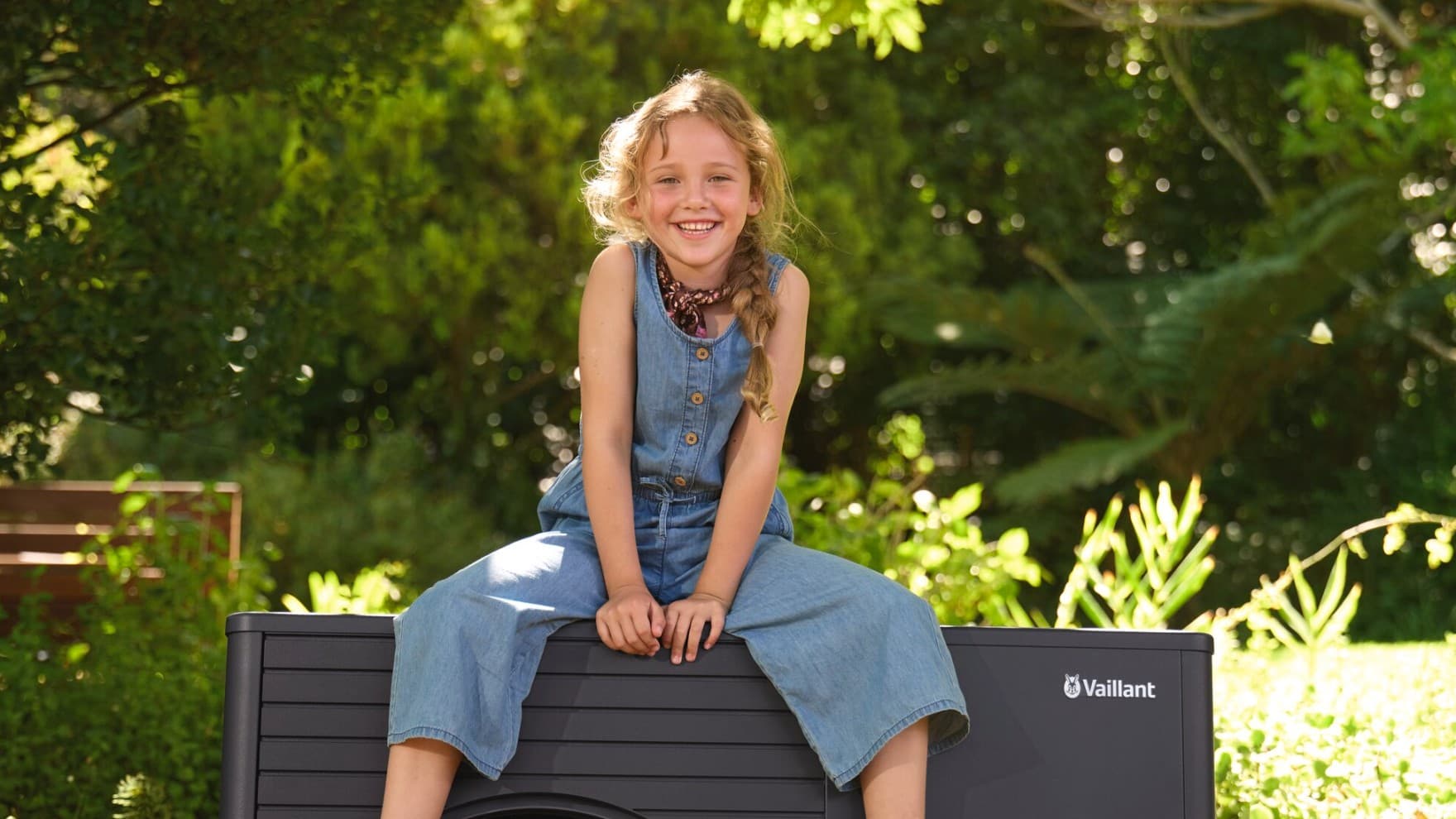 kleines Mädchen sitzt auf Wärmepumpe aroTHERM plus im Garten mit Bäumen im Hintergrund