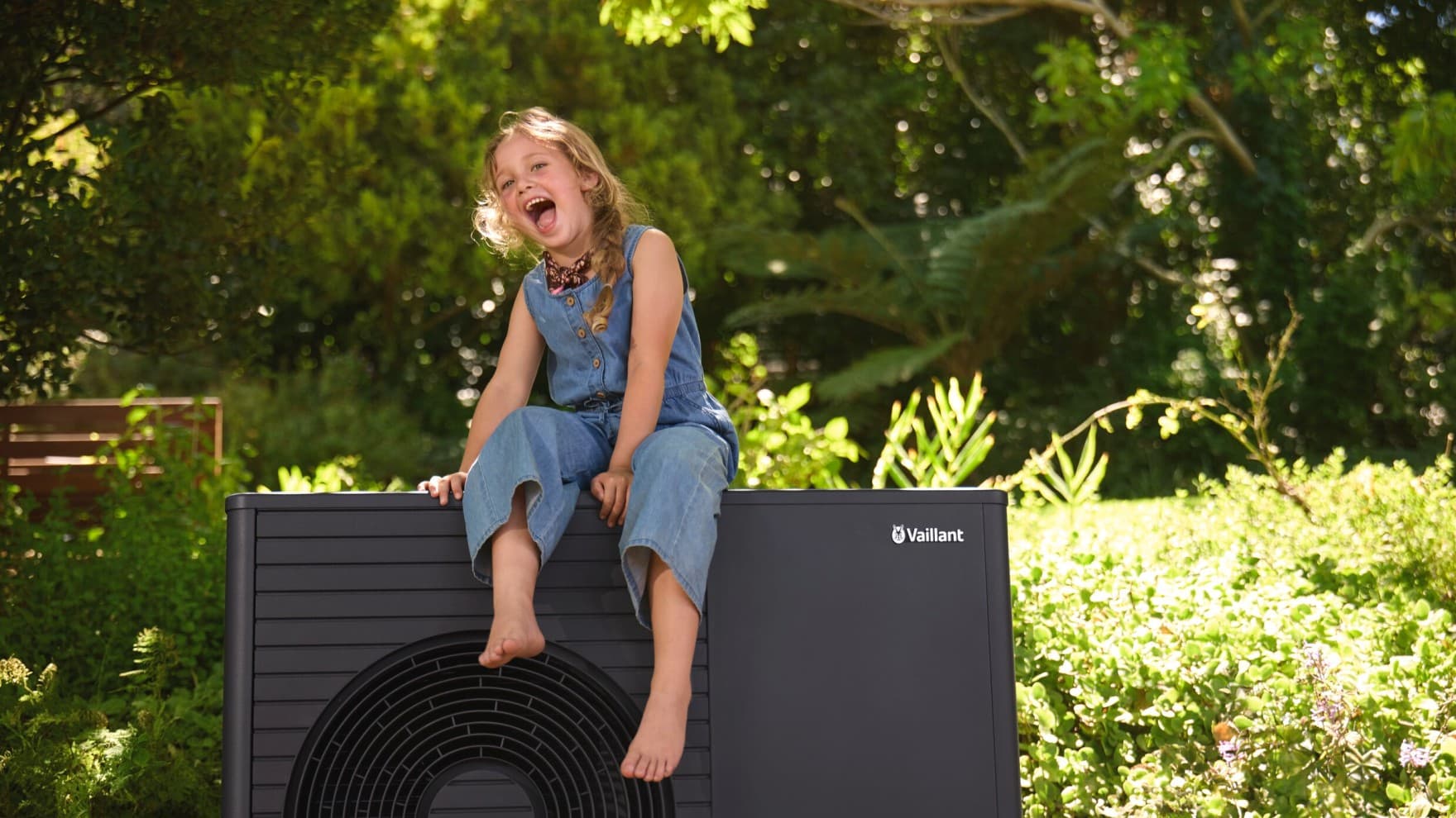 Lachendes, kleines Mädchen sitzt im sommerlichen Garten auf der dunklen Luft/Wasser-Wärmepumpe aroTHERM plus von Vaillant