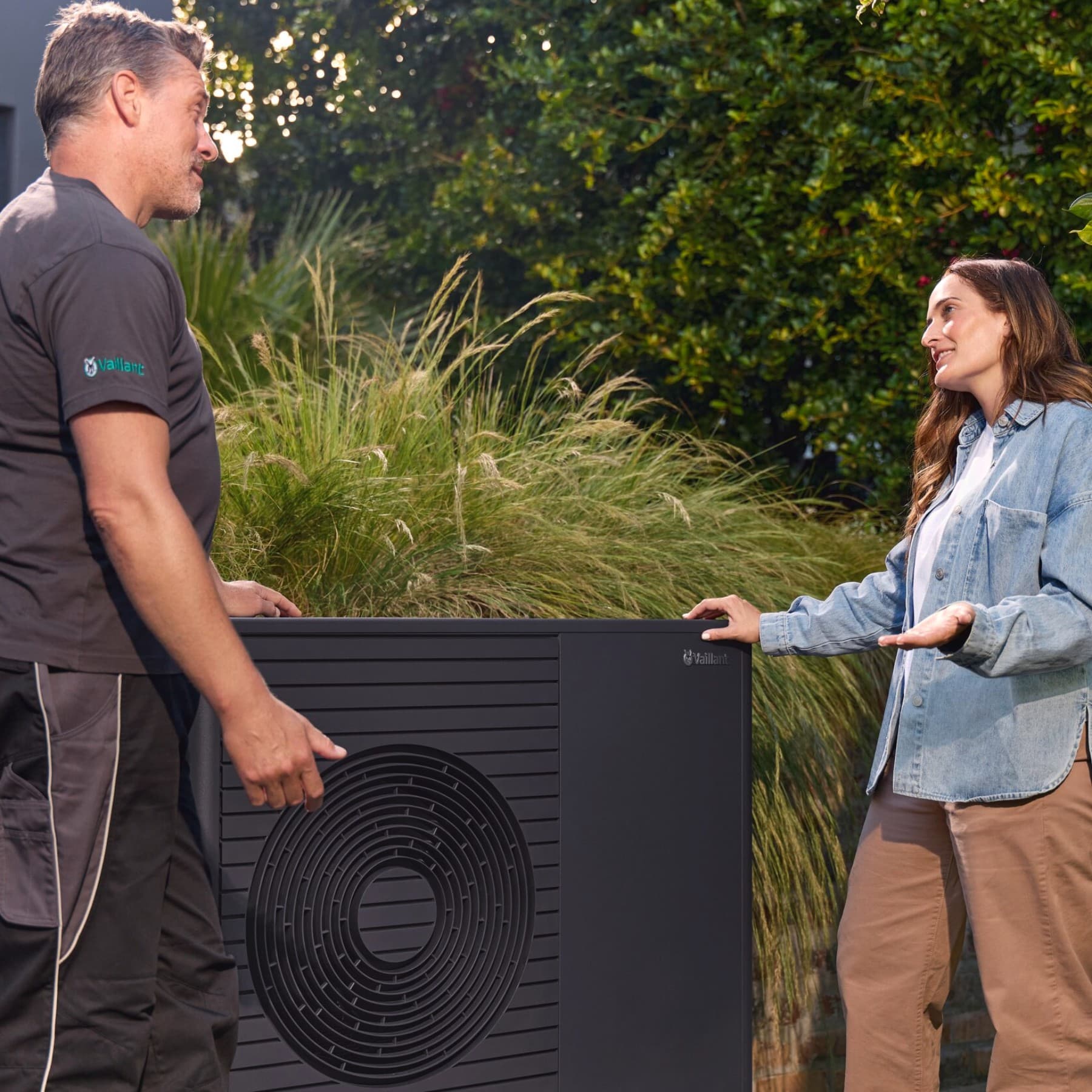 Ein Fachhandwerker im Gespräch mit einer Kundin vor einer Wärmepumpe im Garten