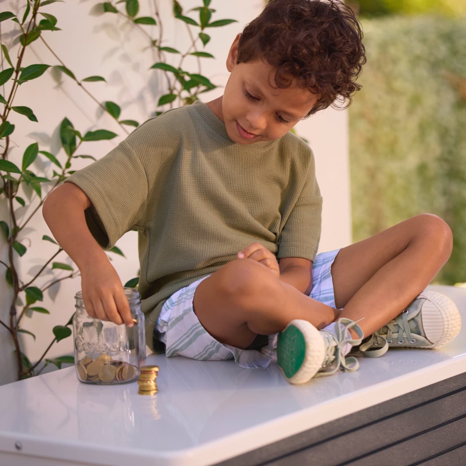 Junge in kurzer Hose sitzt auf einer im sonnigen Garten aufgestellten Vaillant Wärmepumpe und zählt Kleingeld