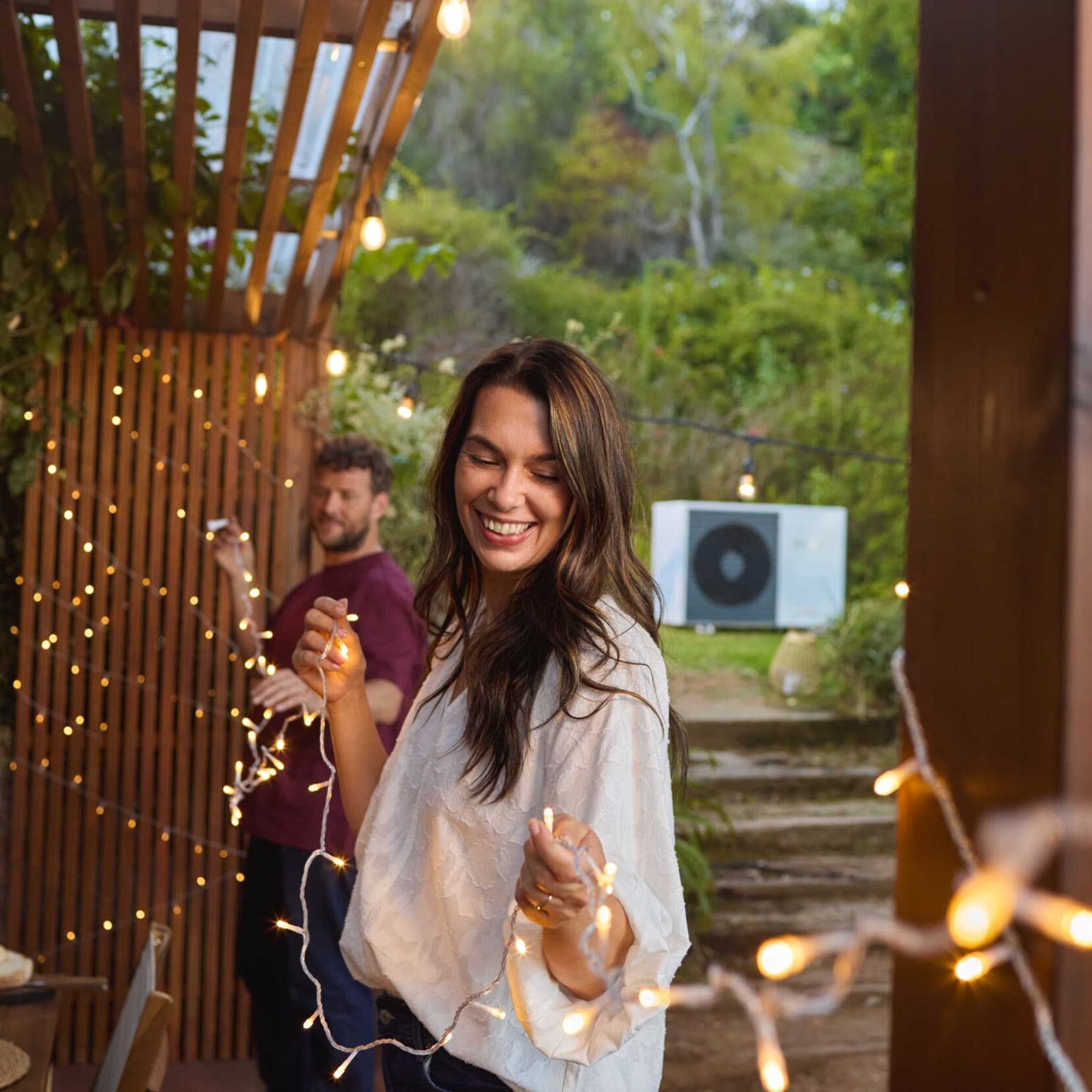 Zwei Personen, die eine Lichterkette aufhängen. Im Hintergrund steht eine Vaillant Wärmepumpe im Garten.