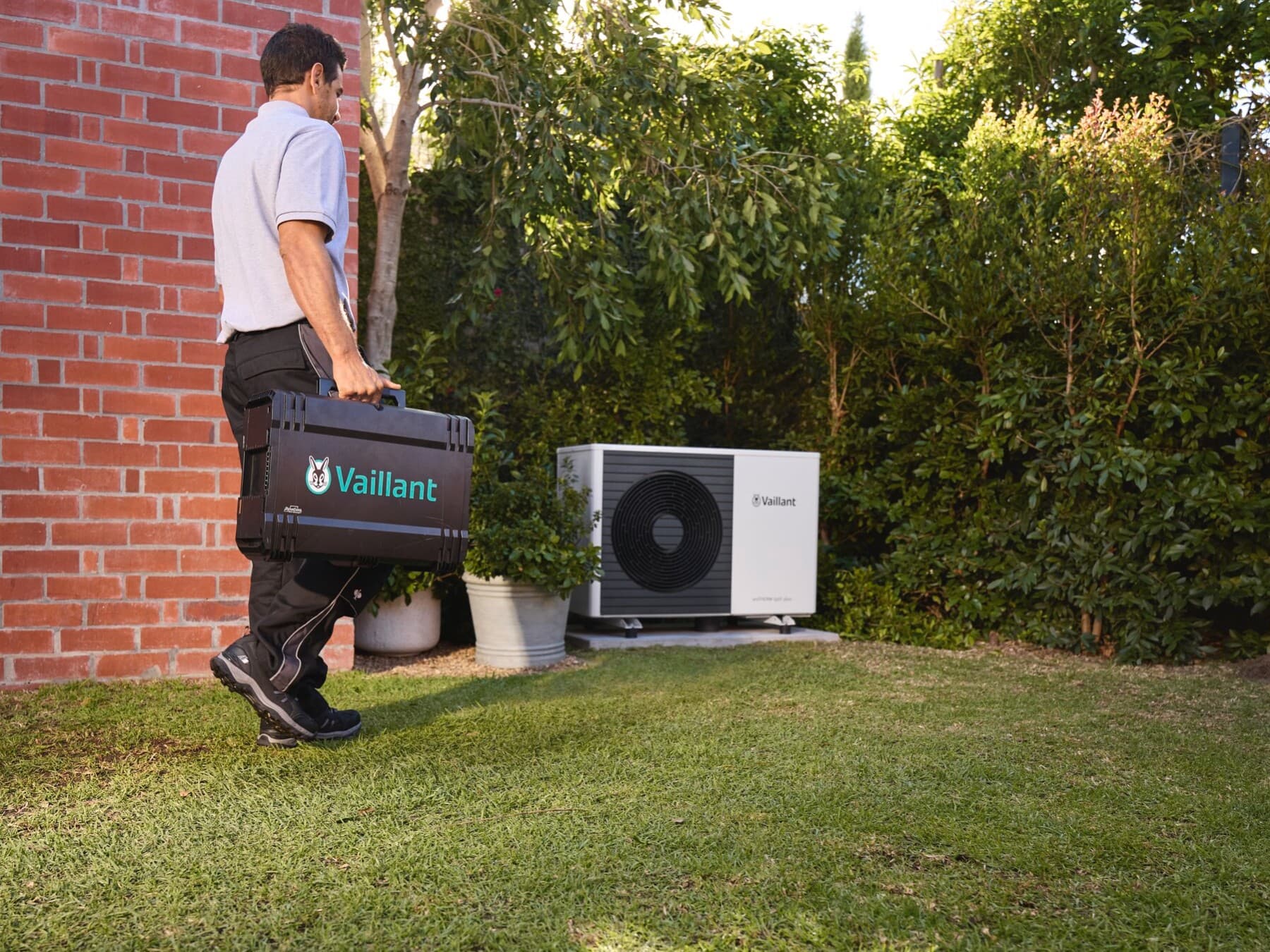 Techniker mit Vaillant-Werkzeugkoffer geht über eine Rasenfläche auf eine im Garten installierte Außeneinheit einer Wärmepumpe zu