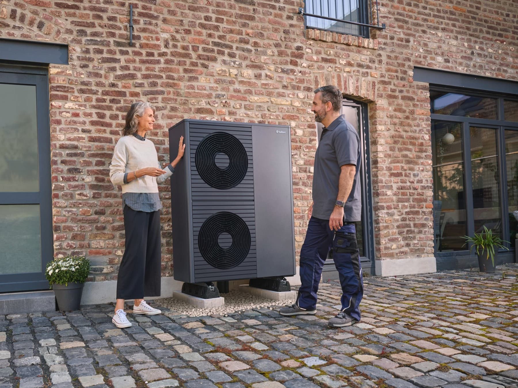 Vor einem Backsteingebäude steht auf Kopfsteinpflaster zwischen zwei Glastüren eine Vaillant Wärmepumpe in der Nähe der Hauswand. Links daneben steht eine Frau, die eine Hand an die Seite der Wärmepumpe legt, rechts von dem Gerät steht ein Vaillant Fachhandwerker. Die beiden schauen sich an.