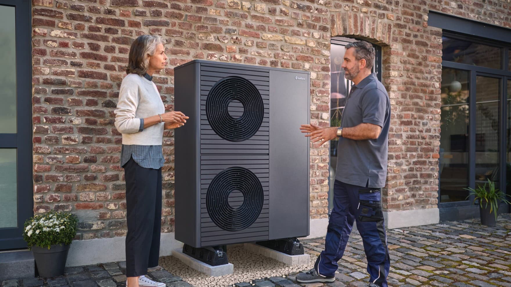 Fachhanwerker und Kundin stehen vor einer neu installierten aroTHERM plus Wärmepumpe vor einem Backsteinhaus