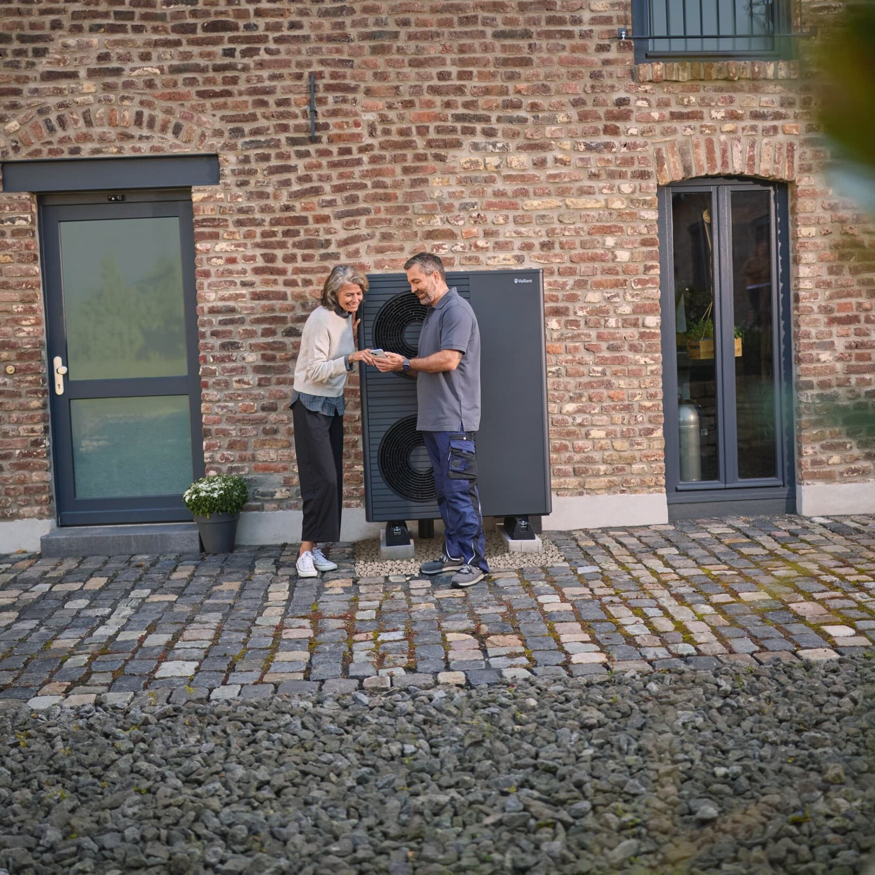 Ein Vaillant Fachhandwerker steht mit einer Kundin vor einer Vaillant Wärmepumpe, die vor einem Backsteingebäude steht und zeigt ihr Unterlagen auf seinem Tablet.