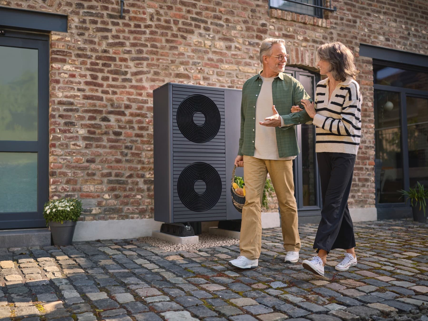 Ein Mann und eine Frau gehen über Kopfsteinpflaster, auf dem hinter ihnen eine Vaillant Wärmepumpe an einer Backsteinhauswand neben einer Glastür steht. Der Mann trägt einen Einkaufskorb, die Frau hat sich bei ihm eingehakt. Die beiden schauen sich an.