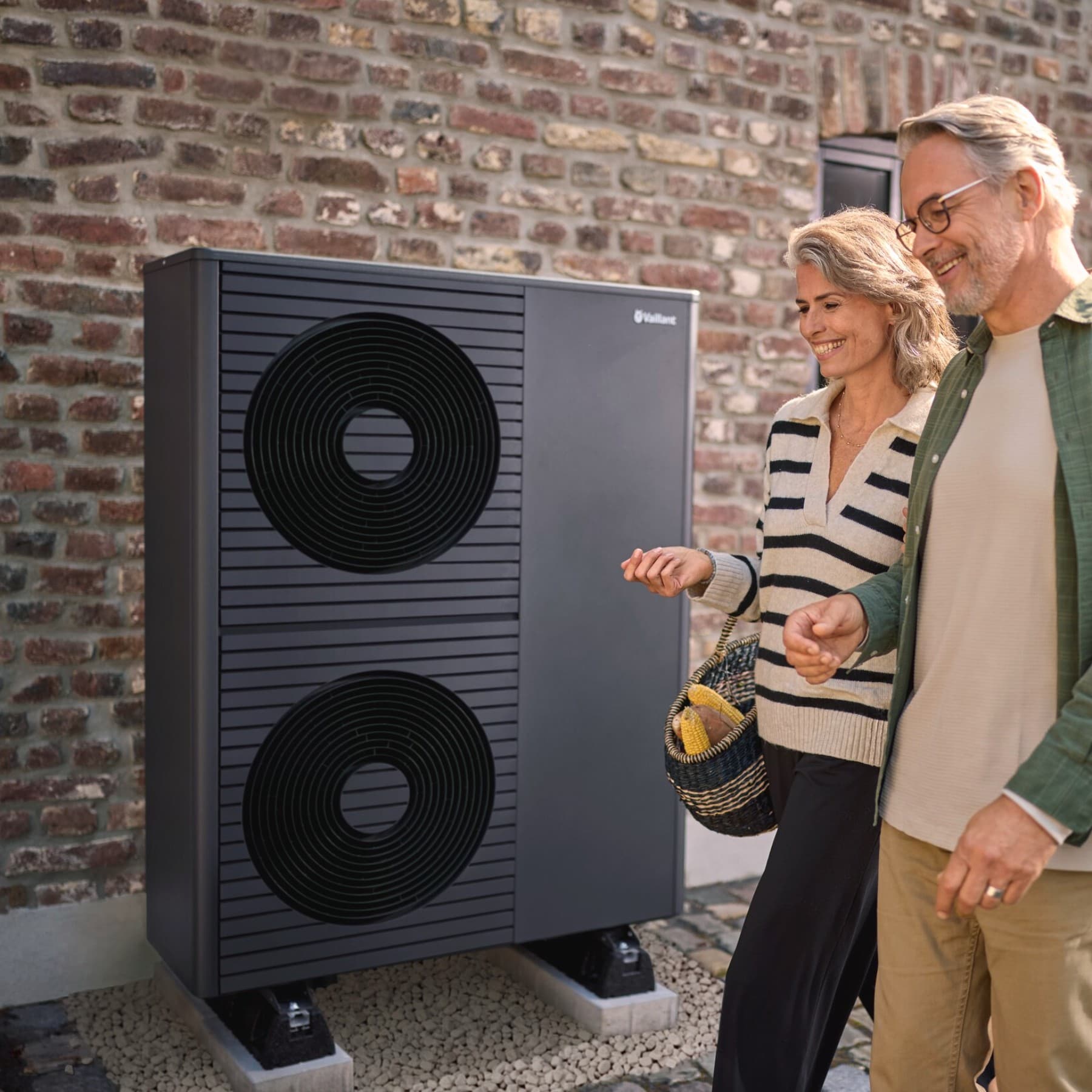 Eine Frau und ein Mann gehen an einem Backsteingebäude vorbei, vor dem eine schwarzgraue Vaillant Wärmepumpe steht.