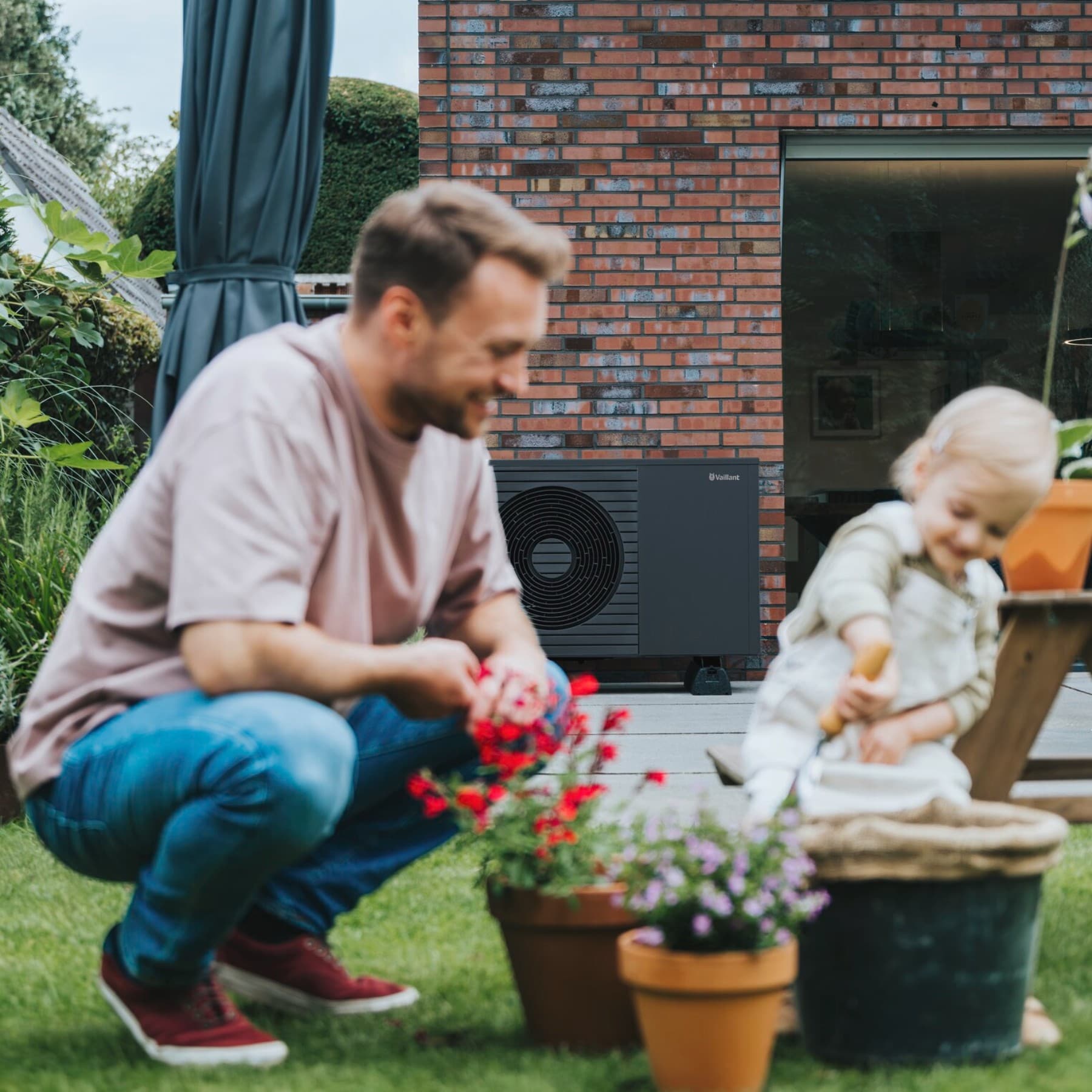 Ein Mann und ein Kind spielen im Garten, sie sind unscharf zu sehen. Der Fokus liegt auf der neuen Vaillant Wärmepumpe aroTHERM plus, die im Hintergrund auf der Terrasse an der Wand des Hauses steht.