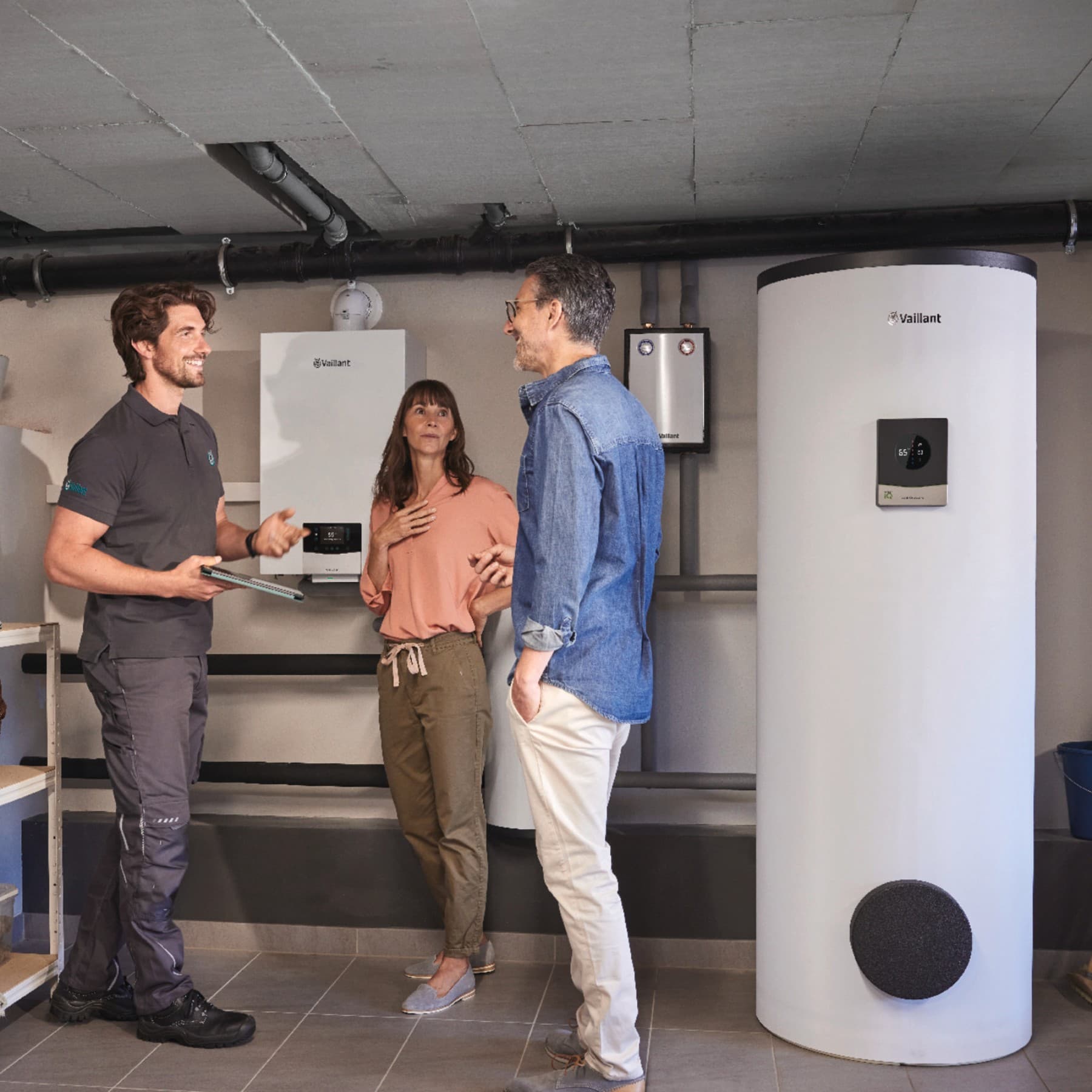 Eine Frau und ein Mann stehen mit einem Vaillant Fachtechniker in einem Heizkeller, in dem eine Vaillant Hybridheizung steht.