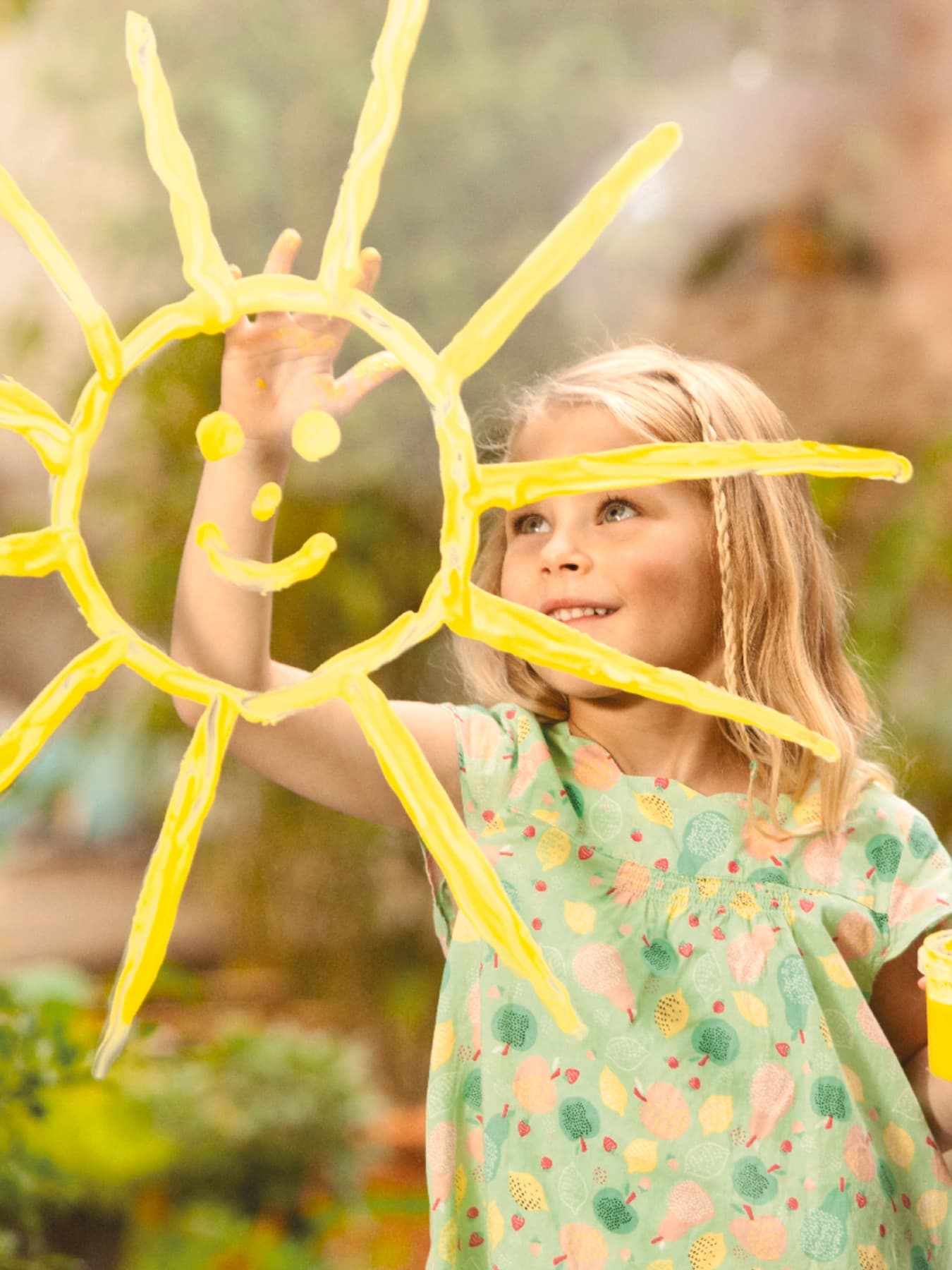 Ein Mädchen im hellgrünen Kleid malt lachend eine Sonne mit gelber Fingerfarbe auf ein Fenster