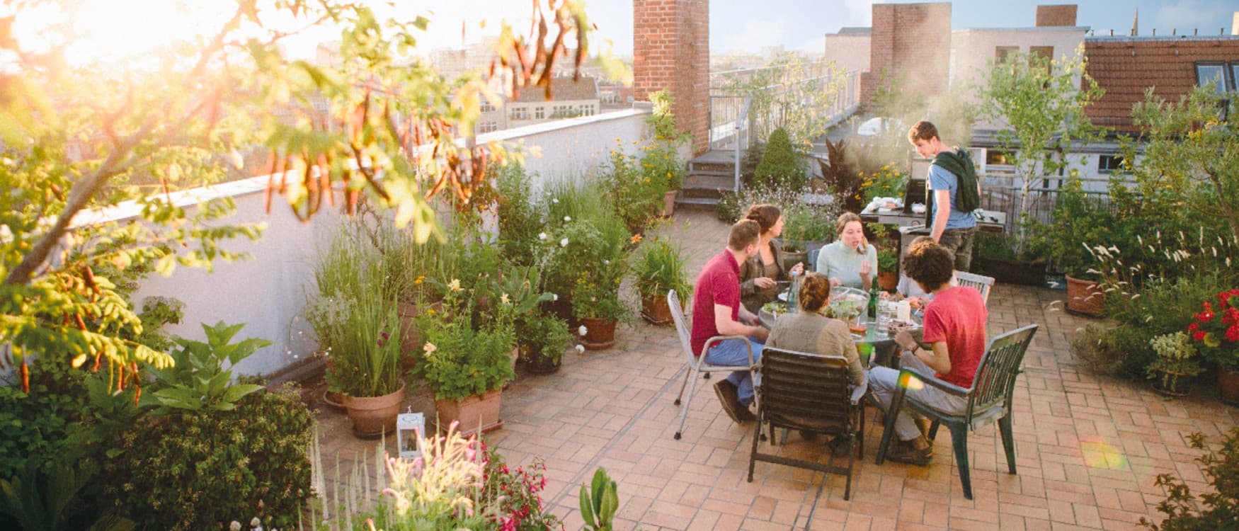 Junge Erwachsene grillen auf einer grün bepflanzten Dachterrasse und genießen den Sommertag zusammen.